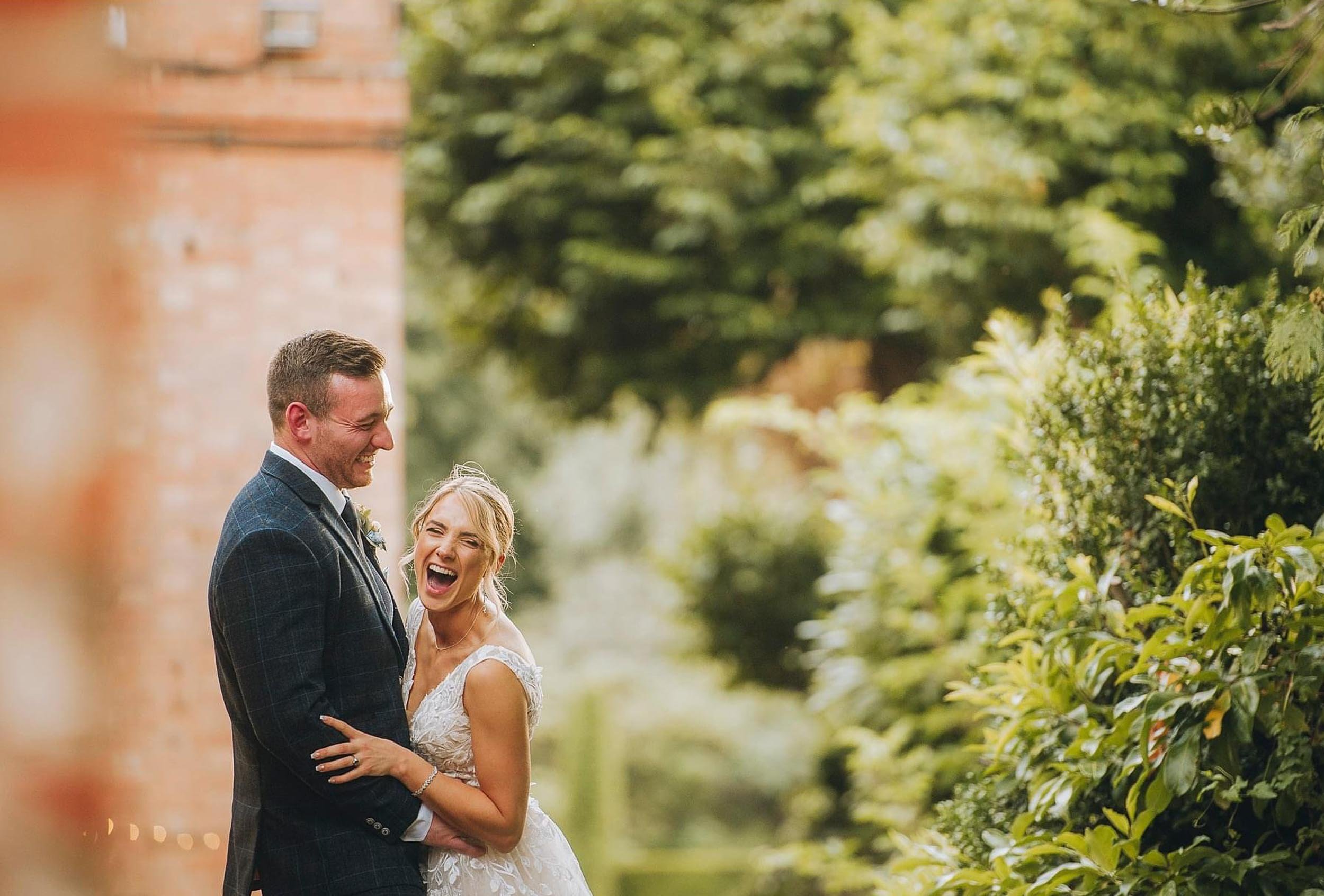 A joyful bride and groom laughing together outdoors surrounded by greenery during their wedding.