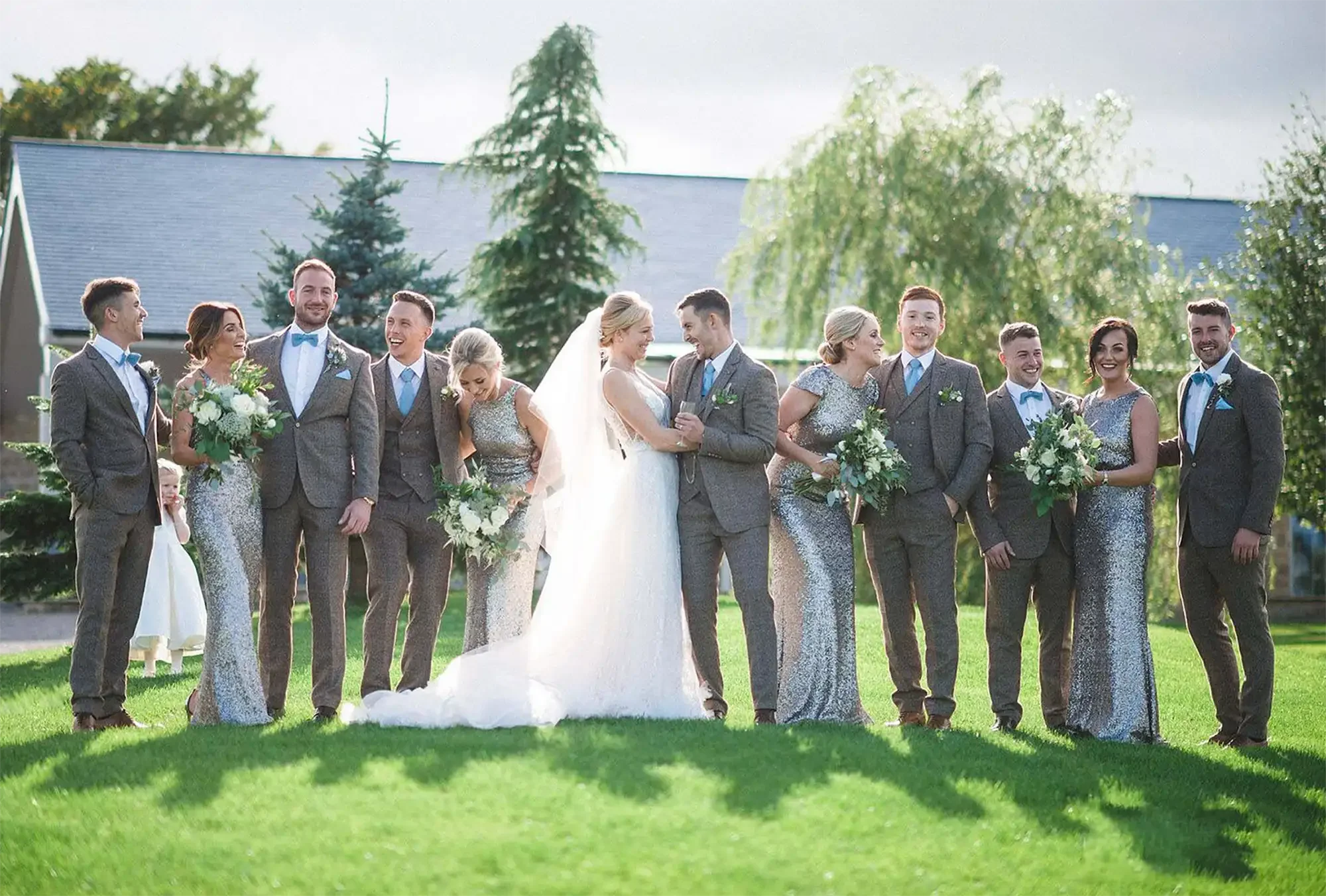 A wedding party stands outdoors on a sunny day with a Yorkshire Wedding Barn in the background. The bride and groom are in the centre, holding hands and smiling. Bridesmaids and groomsmen, stand on either side, holding bouquets of flowers.