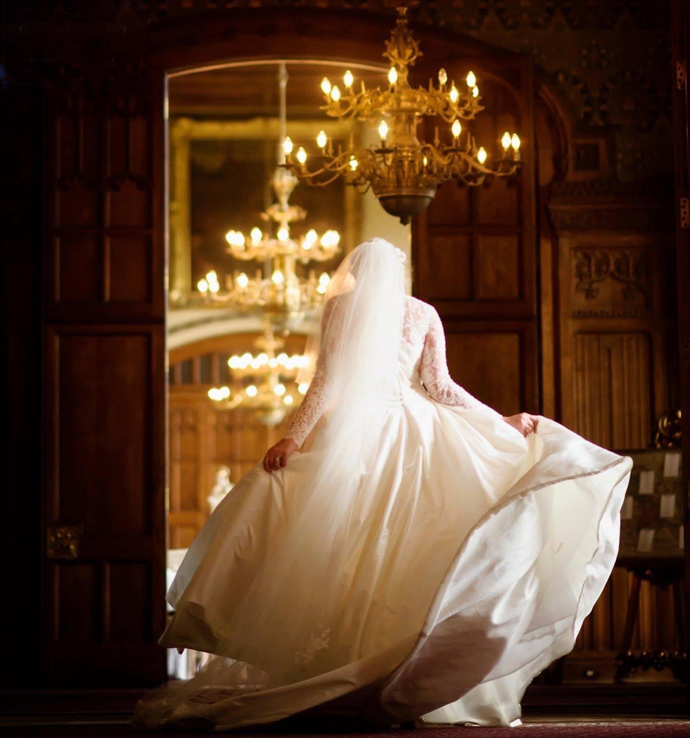 A bride wearing a white wedding dress with long lace sleeves and a veil, standing in an ornately decorated room with wooden walls and a chandelier, holding the skirt of her gown.