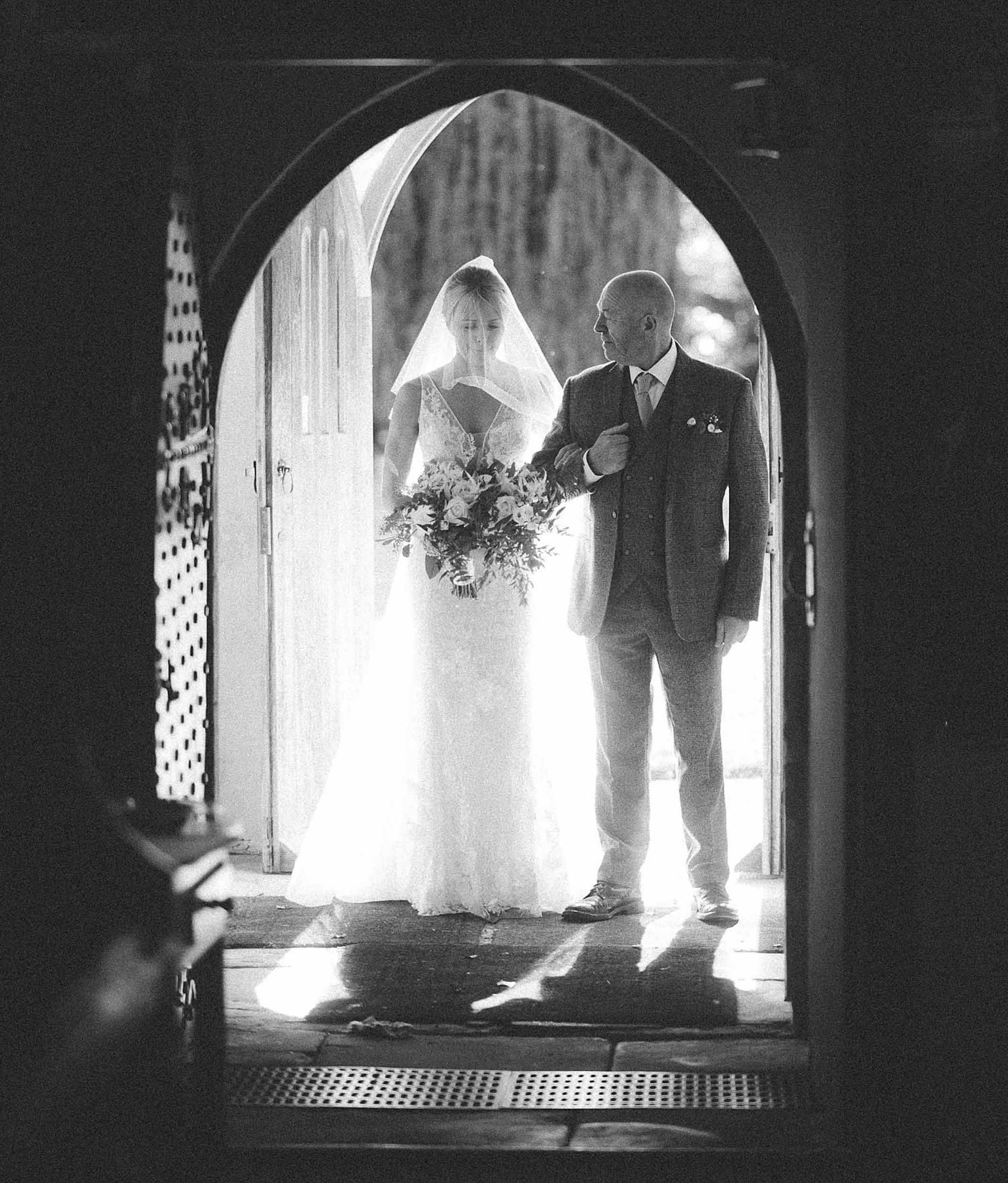 A bride in a wedding dress holding a bouquet, walking down an aisle with an older man, possibly her father, inside a church or chapel
