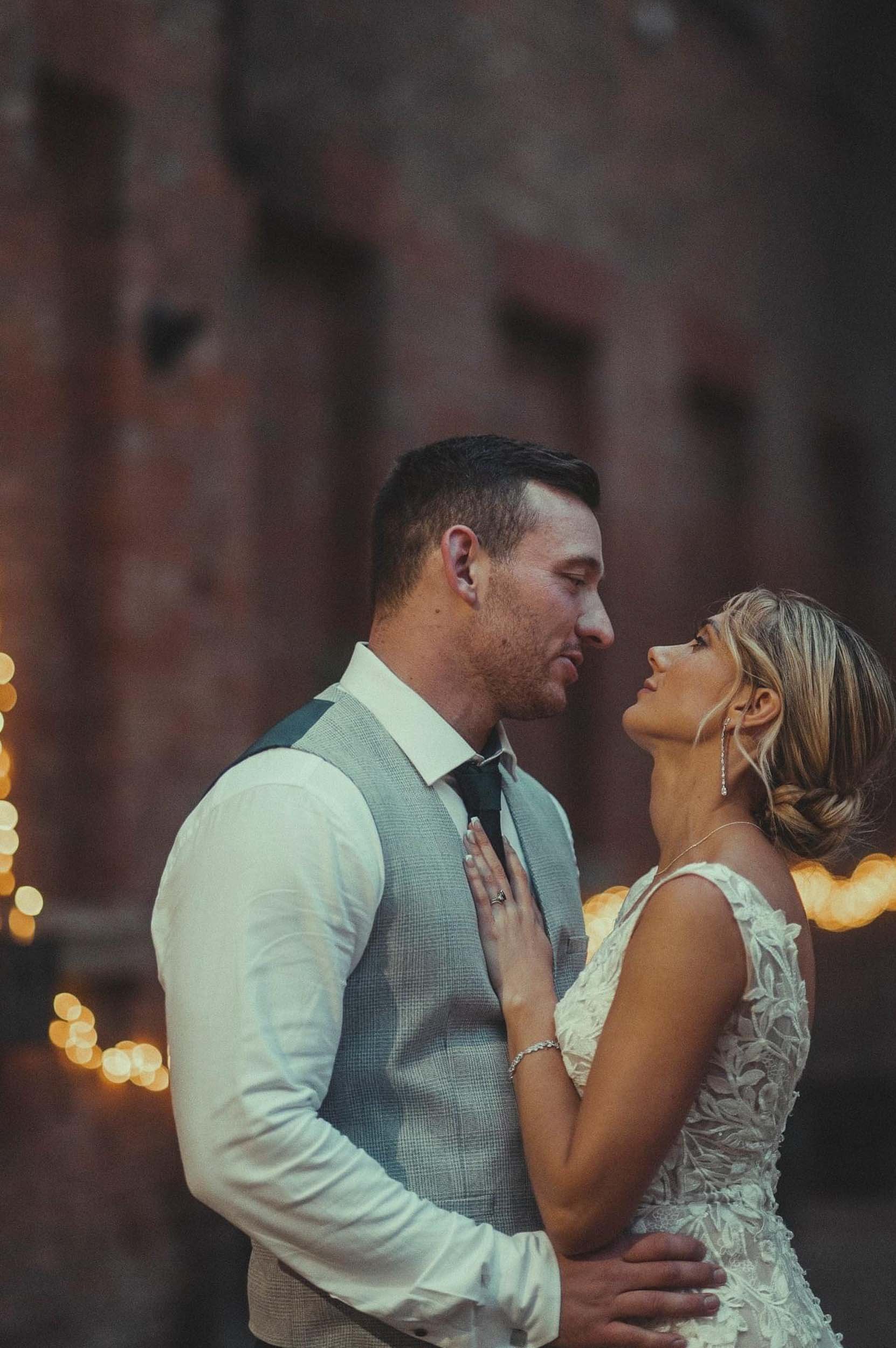 A couple in wedding attire sharing a romantic moment, looking into each other's eyes, with blurred string lights in the background.