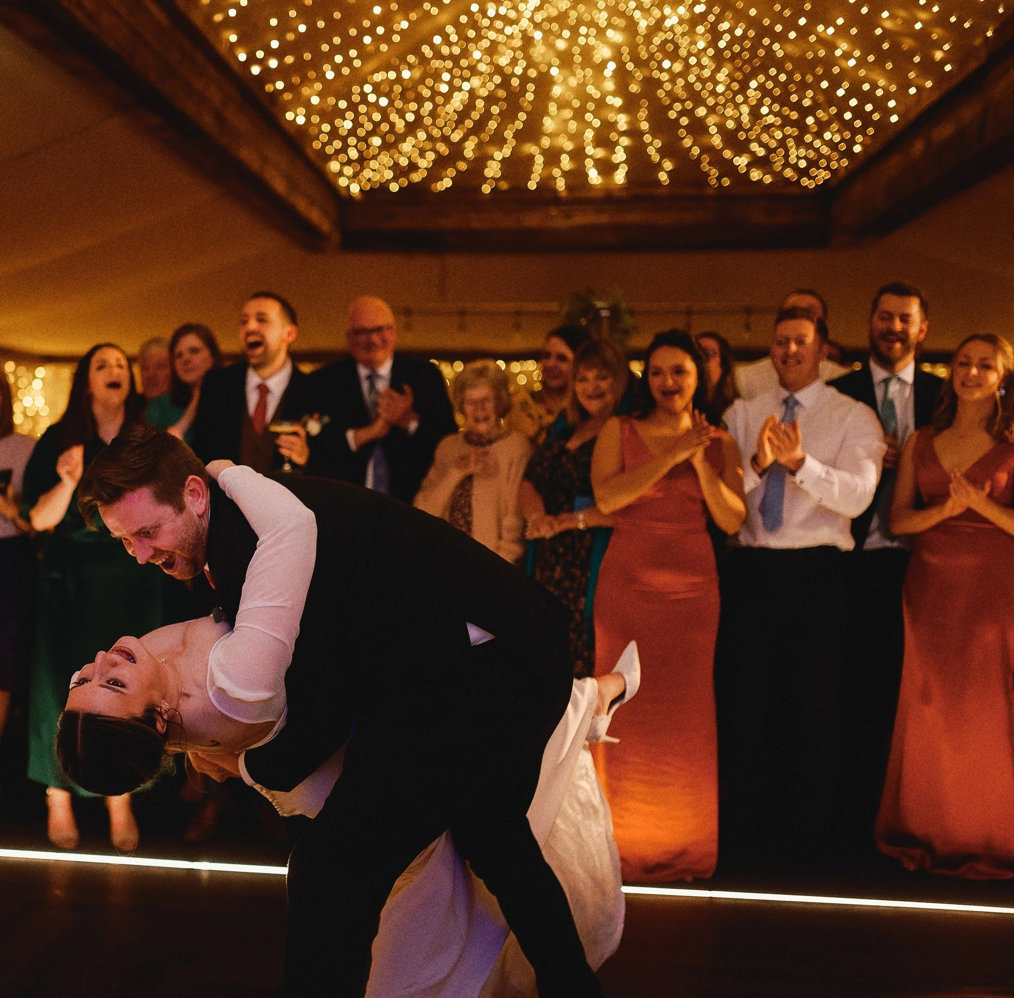 A newlywed couple sharing their first dance at a wedding reception, surrounded by friends and family who are smiling and clapping under a ceiling decorated with string lights.