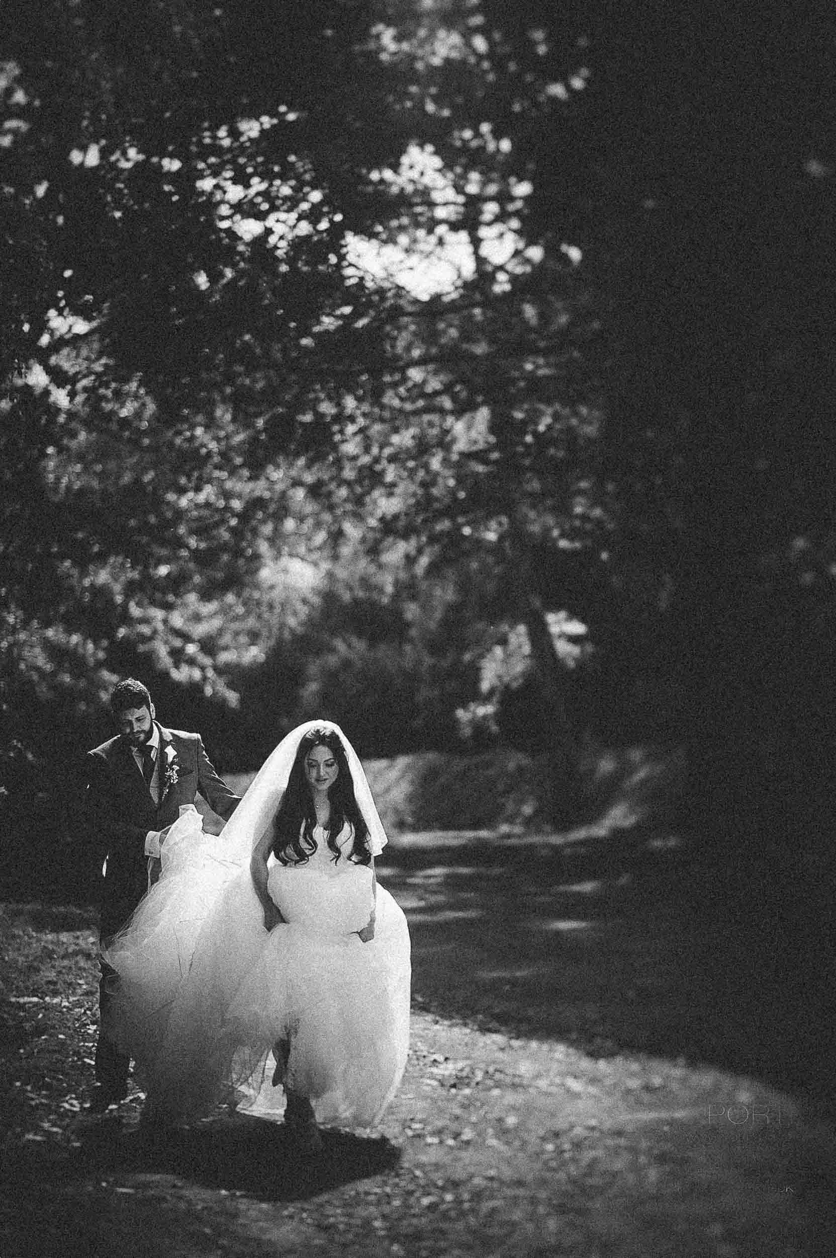 A black and white photograph of a bride and groom walking outdoors at night, with trees in the background. The bride is holding her wedding dress, and the groom is assisting her.