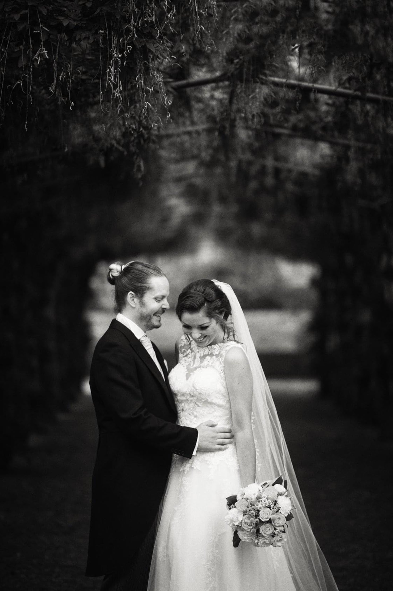 A black and white photo of a bride and groom smiling and embracing outdoors under a natural archway formed by trees, with the bride holding a bouquet of flowers.