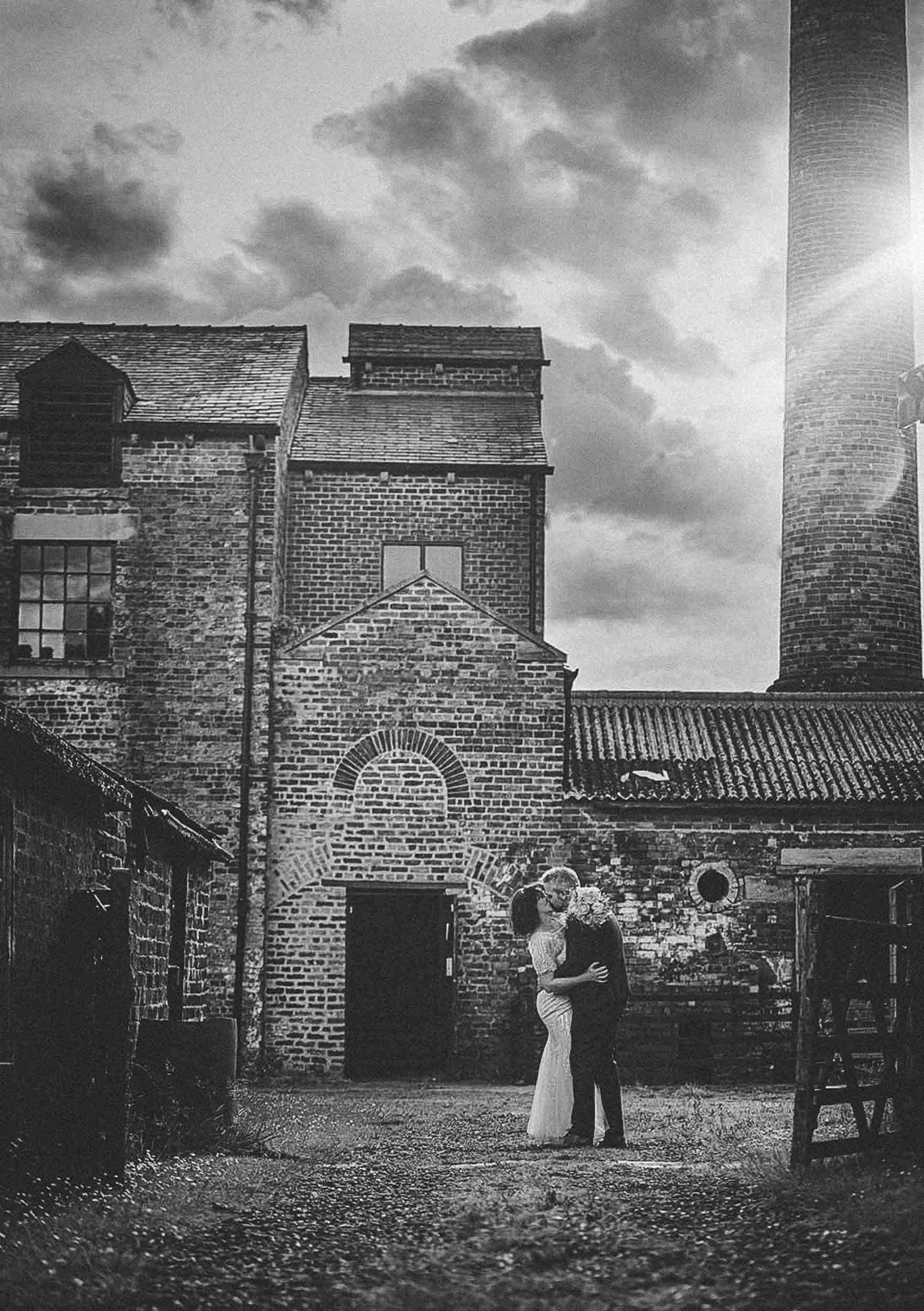 A couple in wedding attire embraces and kisses in an alleyway with old brick buildings, a tall chimney, and a cloudy sky in the background.