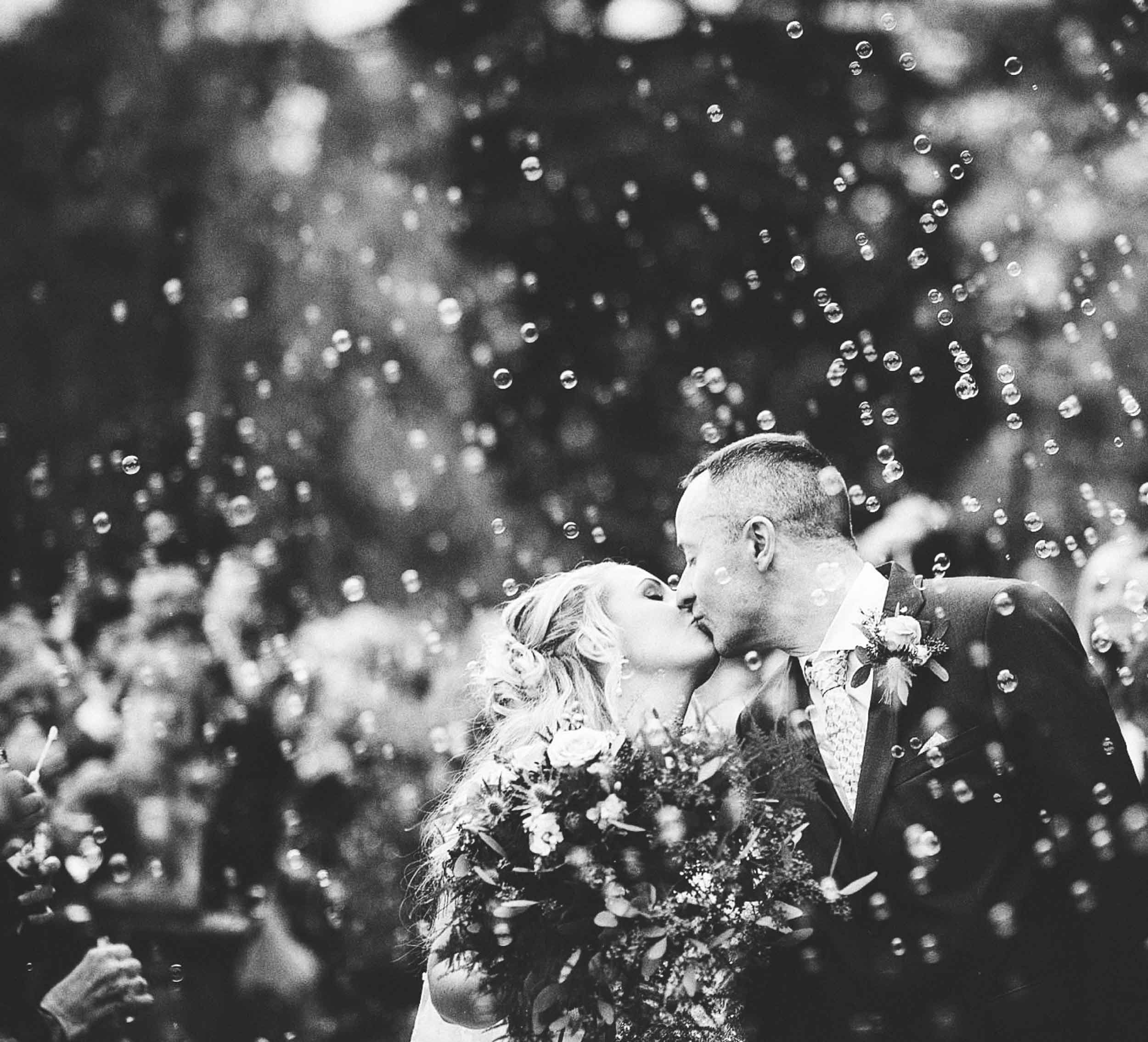 A black and white photo of a bride and groom kissing at their wedding, surrounded by guests and falling confetti or bubbles.