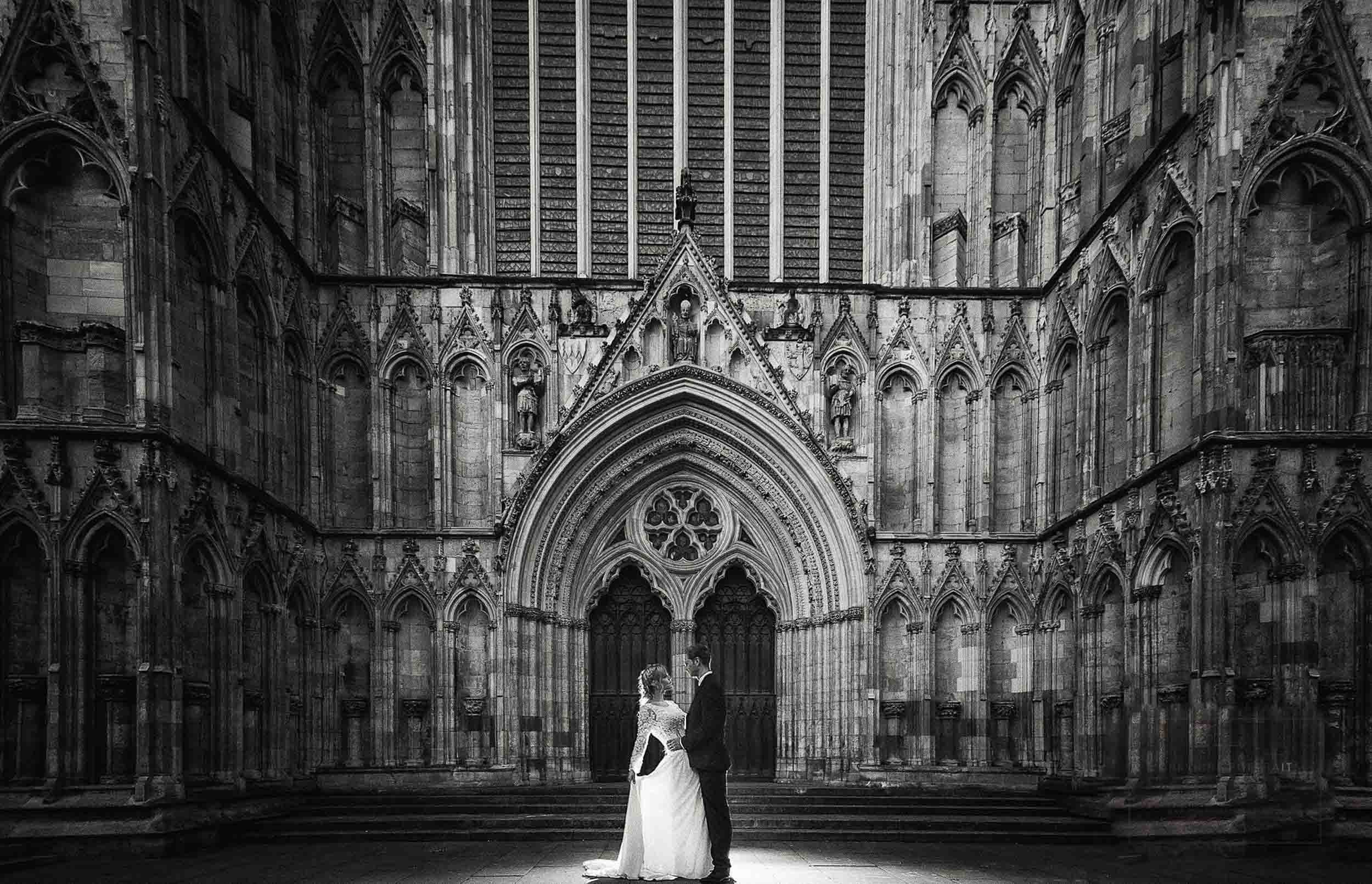 A bride and groom stand facing each other in front of a large gothic cathedral entrance with detailed stone carving.