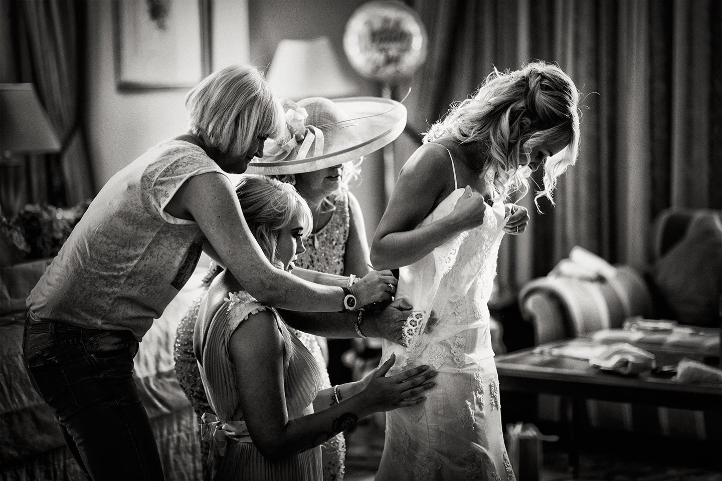 Women helping a bride put on her wedding dress in a room with curtains and furniture.