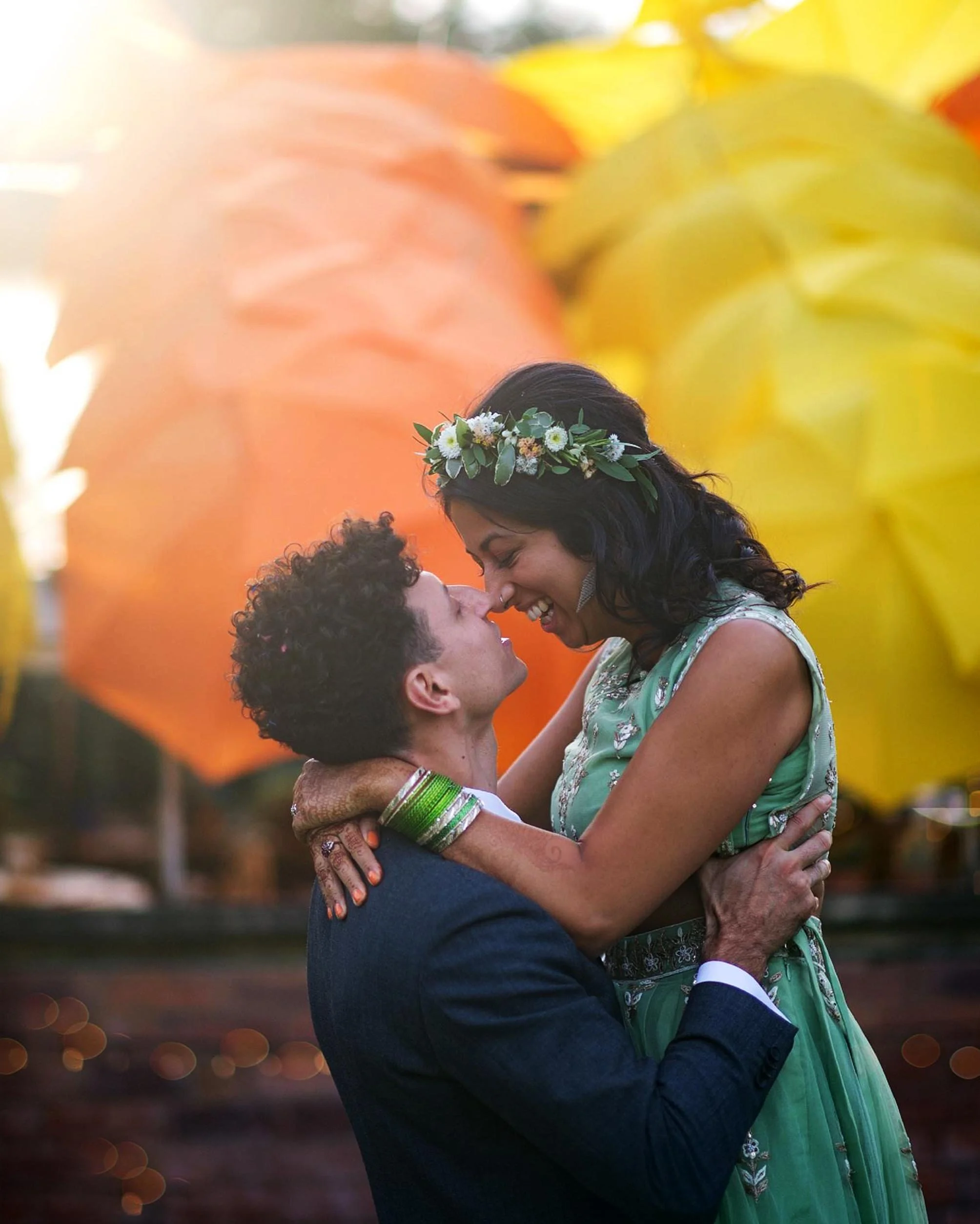 A couple is embracing and about to kiss at sunset, with large colorful umbrellas in the background.
