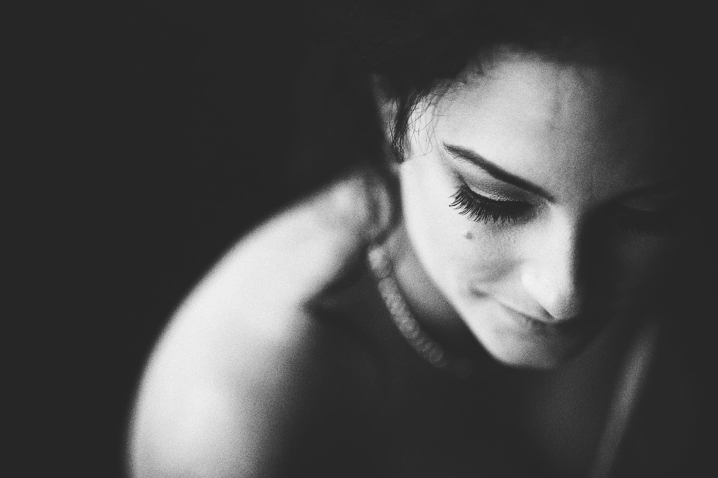 Black and white close-up portrait of a woman with eyes closed, smiling softly, dark background, wearing a necklace.
