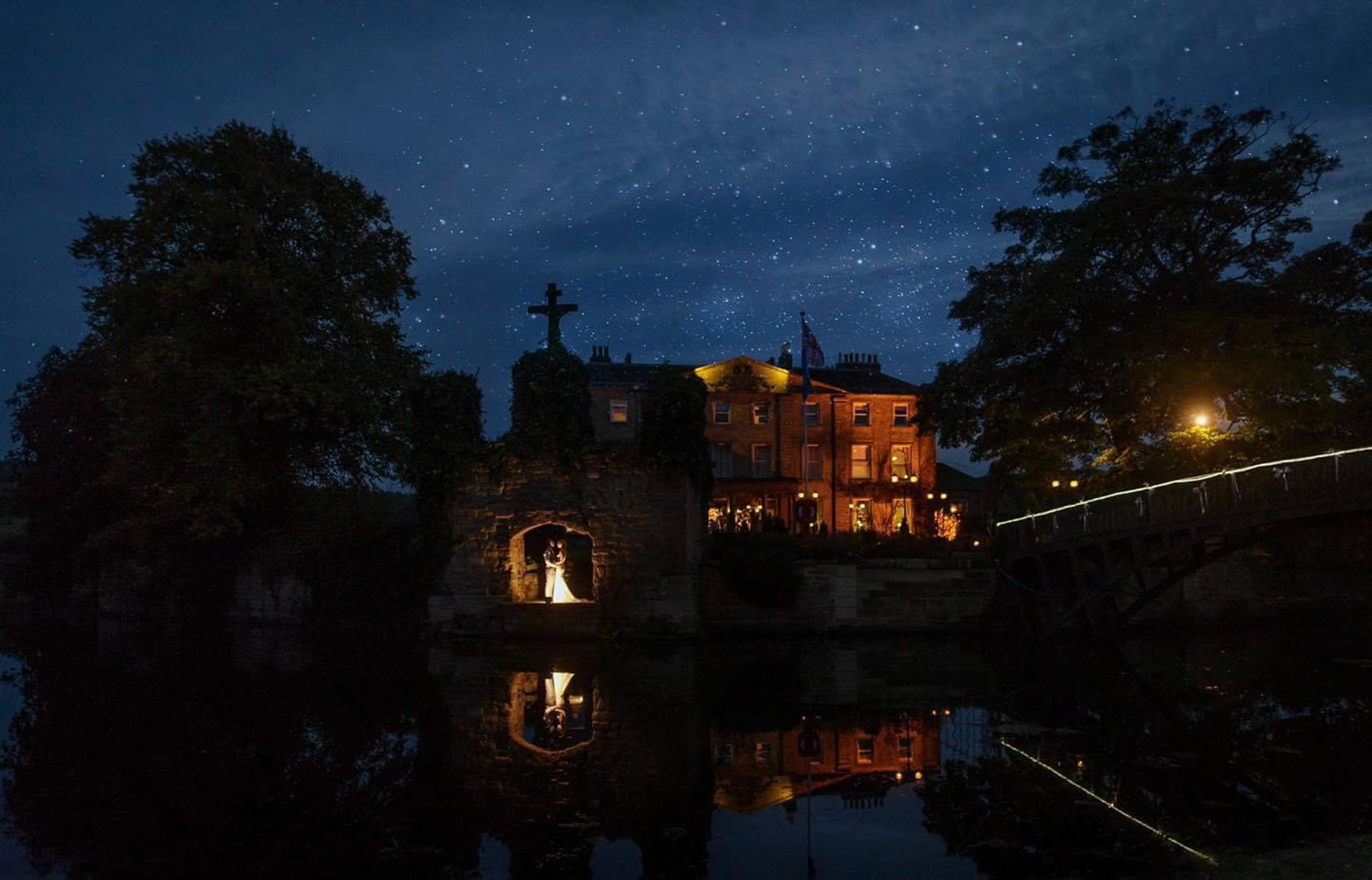 Night view of a historic building with a large cross on top, illuminated by warm lights, reflecting in a river below. Starry sky overhead with trees on each side and a small illuminated bridge on the right.