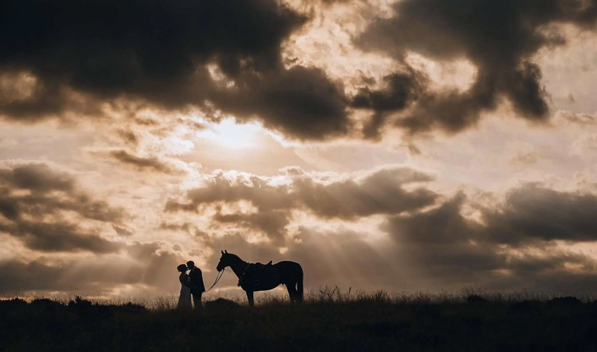 Silhouette of a couple kissing while holding a leash attached to a horse, with a dramatic cloud-filled sky and setting sun in the background.