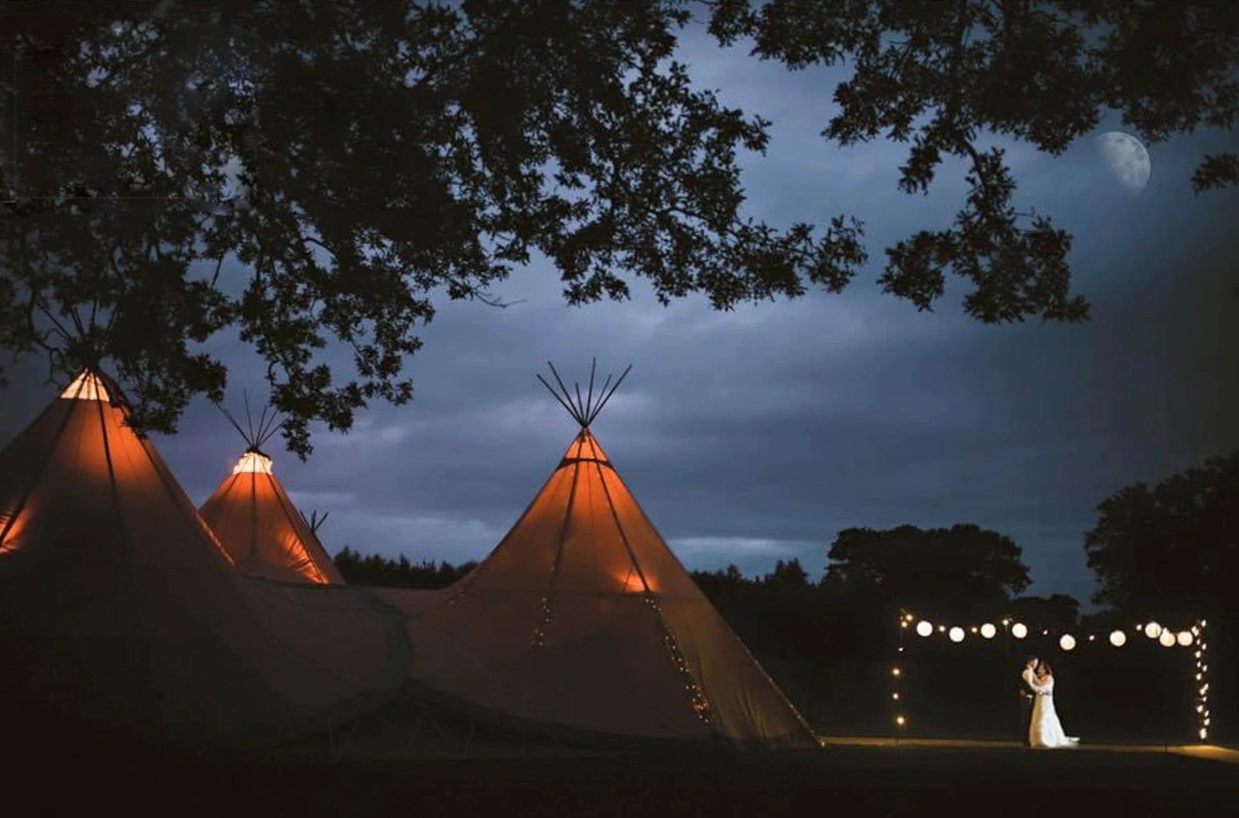 A couple dancing at night outside under string lights near teepees, with a full moon and trees in the background.