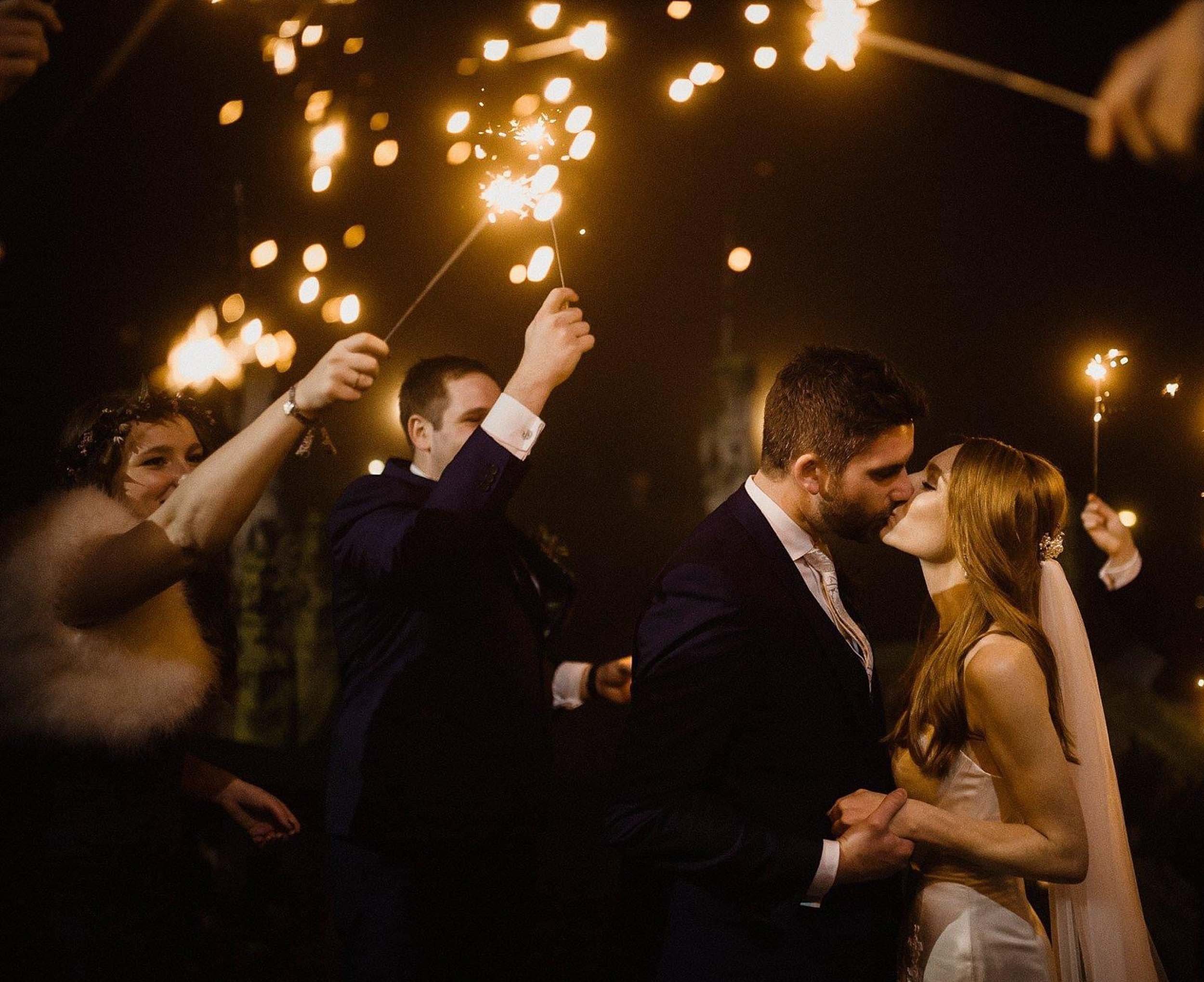 A bride and groom kiss at night, celebrating their wedding as friends hold sparklers overhead.