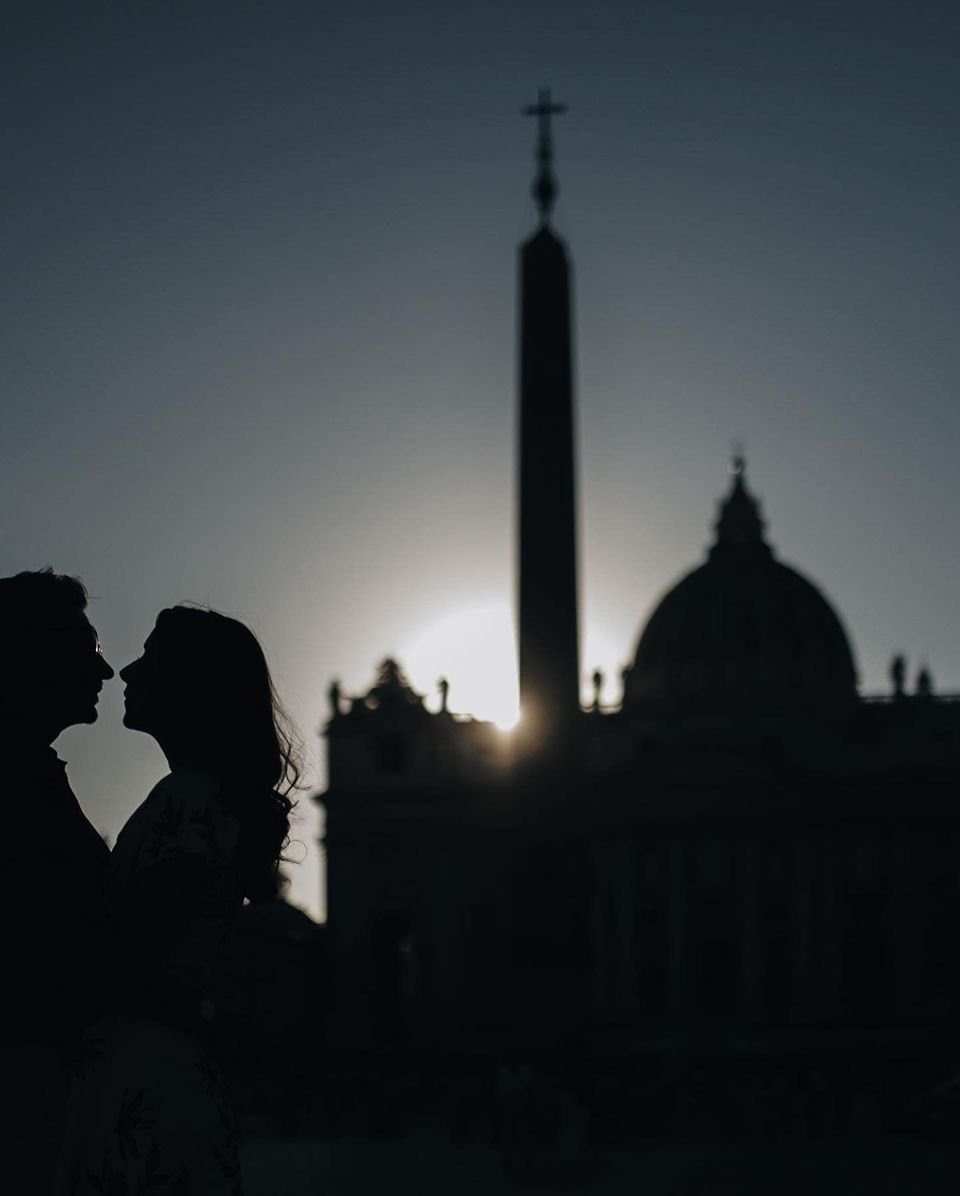 Silhouette of a couple facing each other at sunset with historic buildings in the background, including a dome and an obelisk.