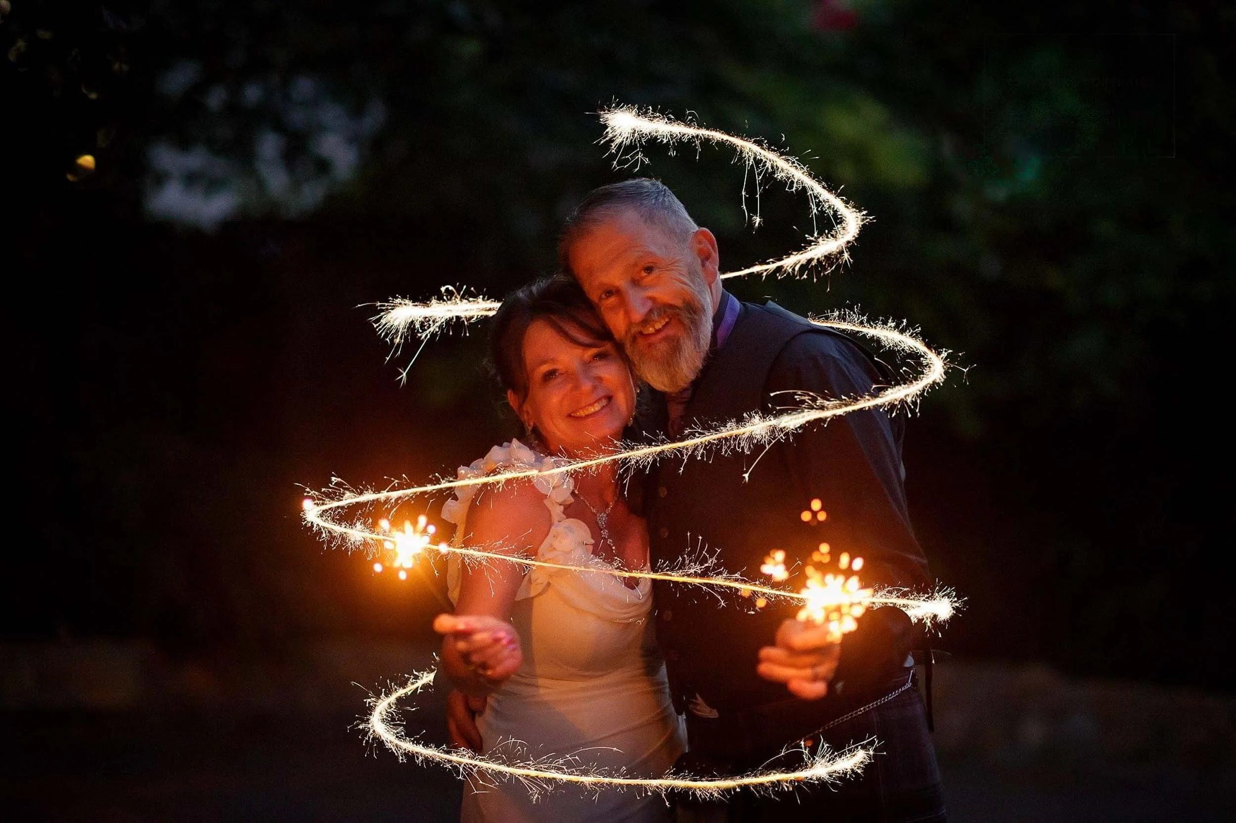 A couple smiling and embracing, holding sparklers at night, with glowing sparklers creating a spiral light pattern around them.