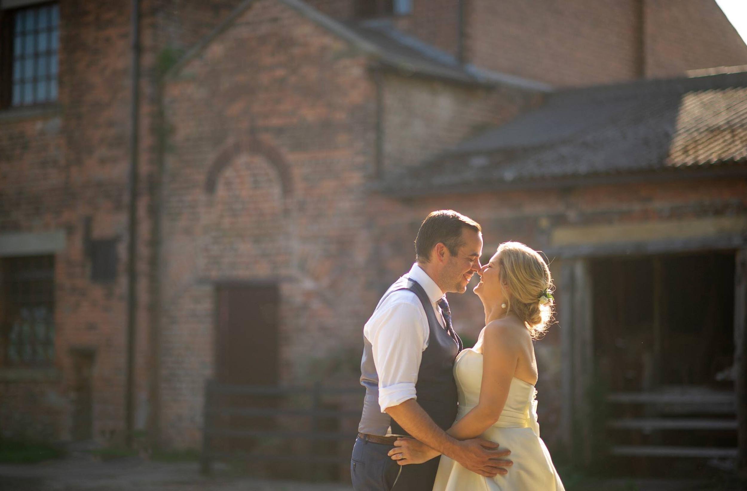 A couple dressed in wedding attire sharing an intimate moment outdoors near a brick building.