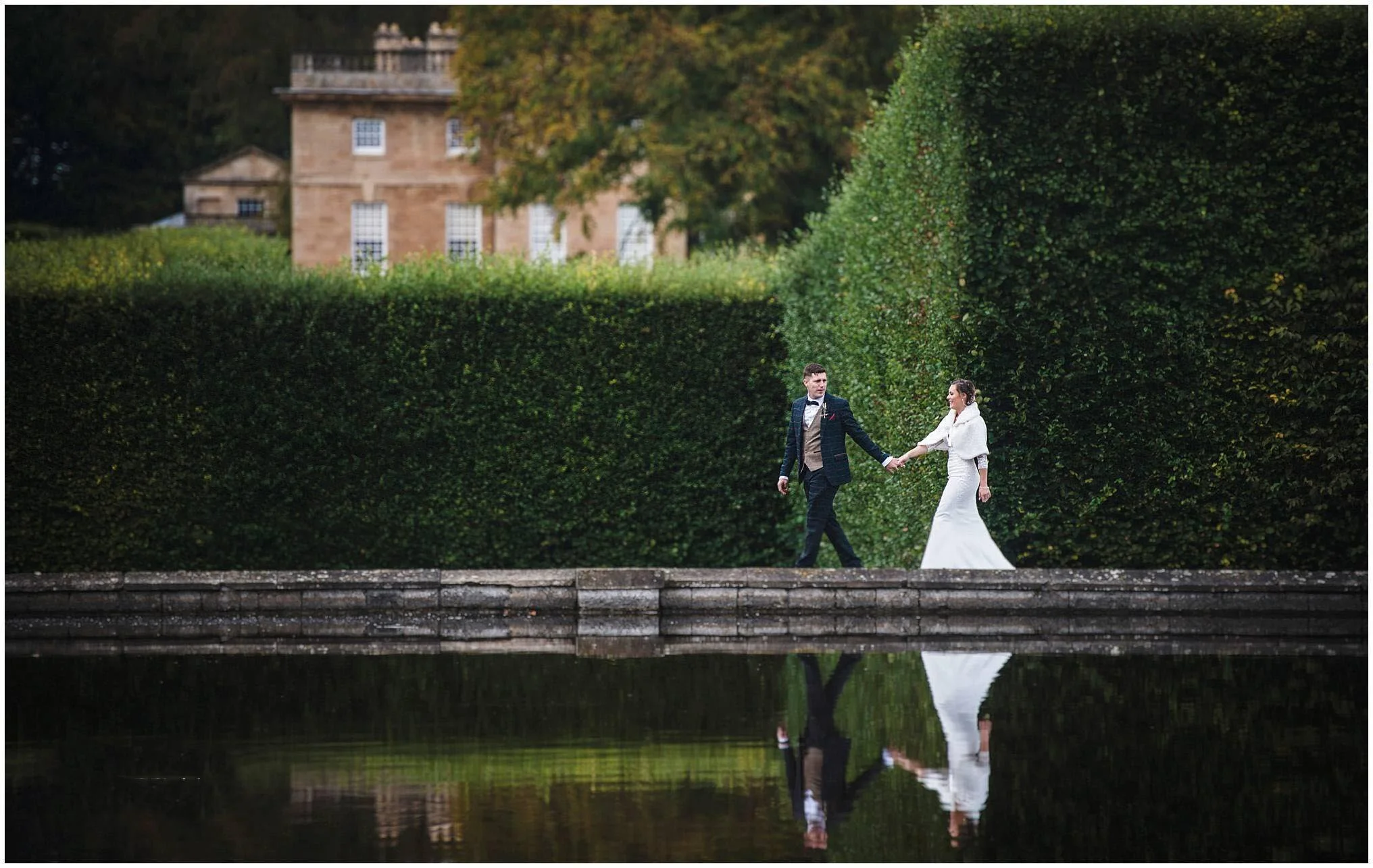 A bride and groom walk hand in hand along a stone pathway beside a pond, with lush green hedges and a historic building in the background.