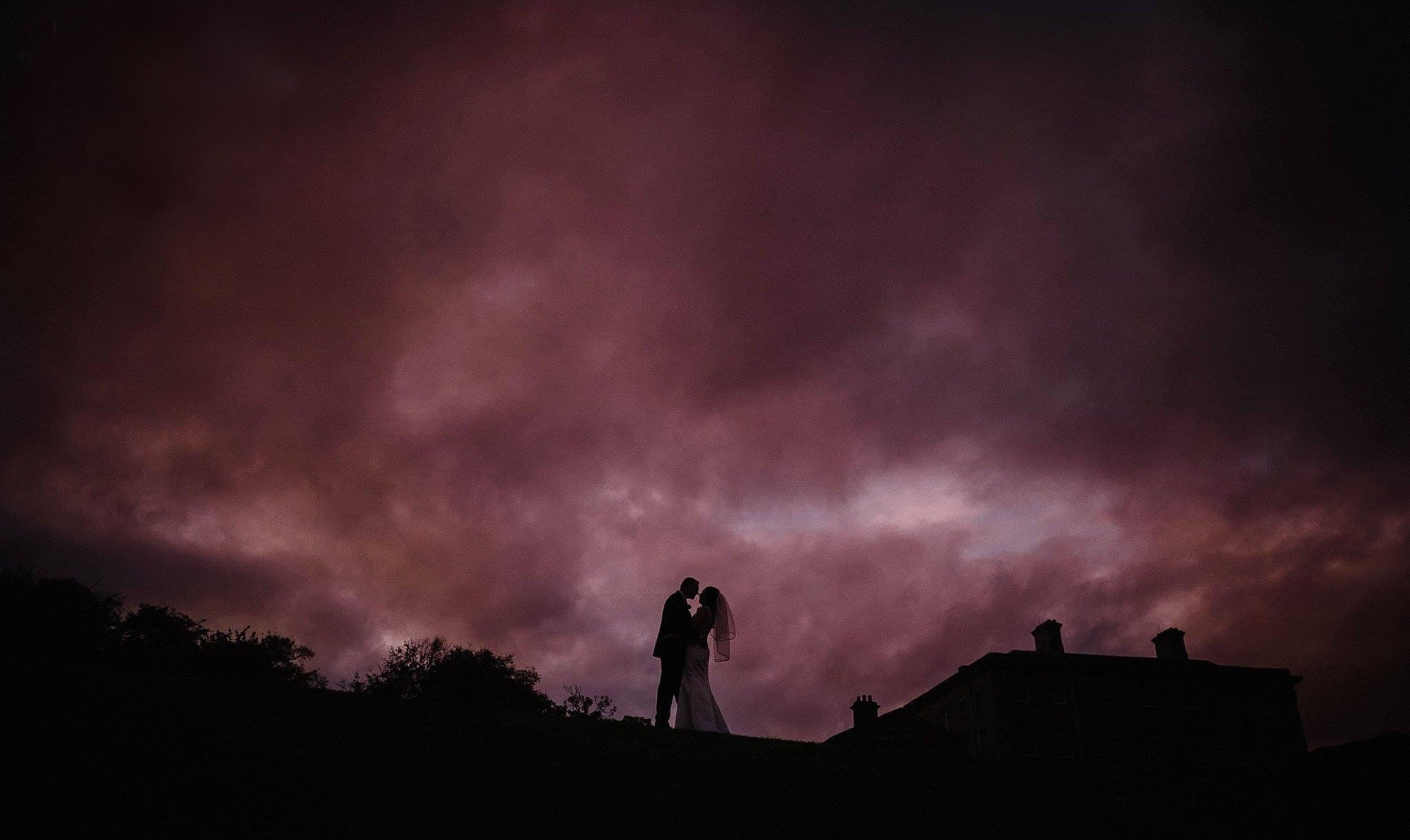 Silhouetted couple, possibly newlyweds, standing close and touching foreheads against a dark, cloudy sky at dusk, with a house silhouette in the foreground.