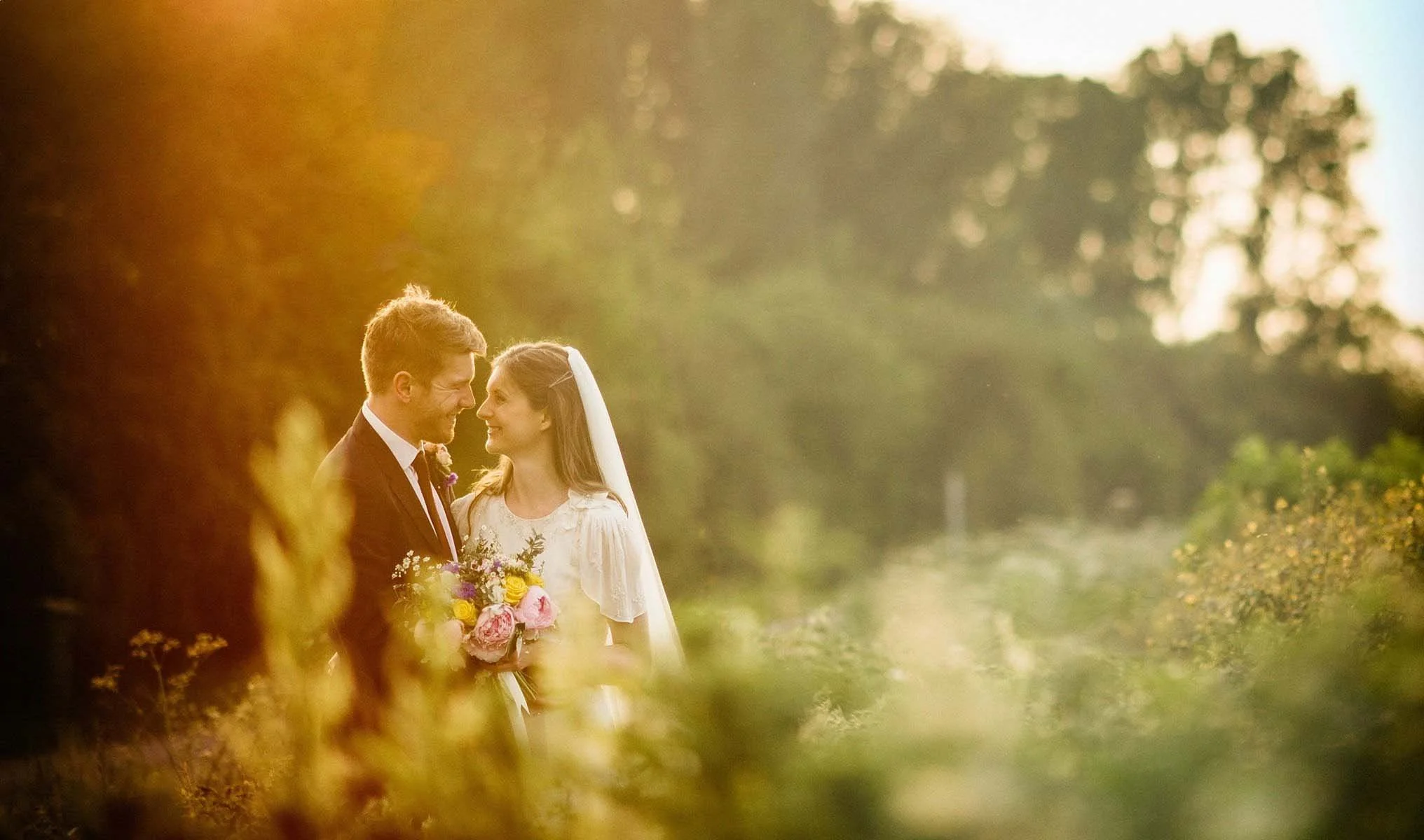 A bride and groom on their wedding day, standing closely and gazing at each other, with the bride holding a bouquet of flowers, outdoors during sunset in a natural setting.