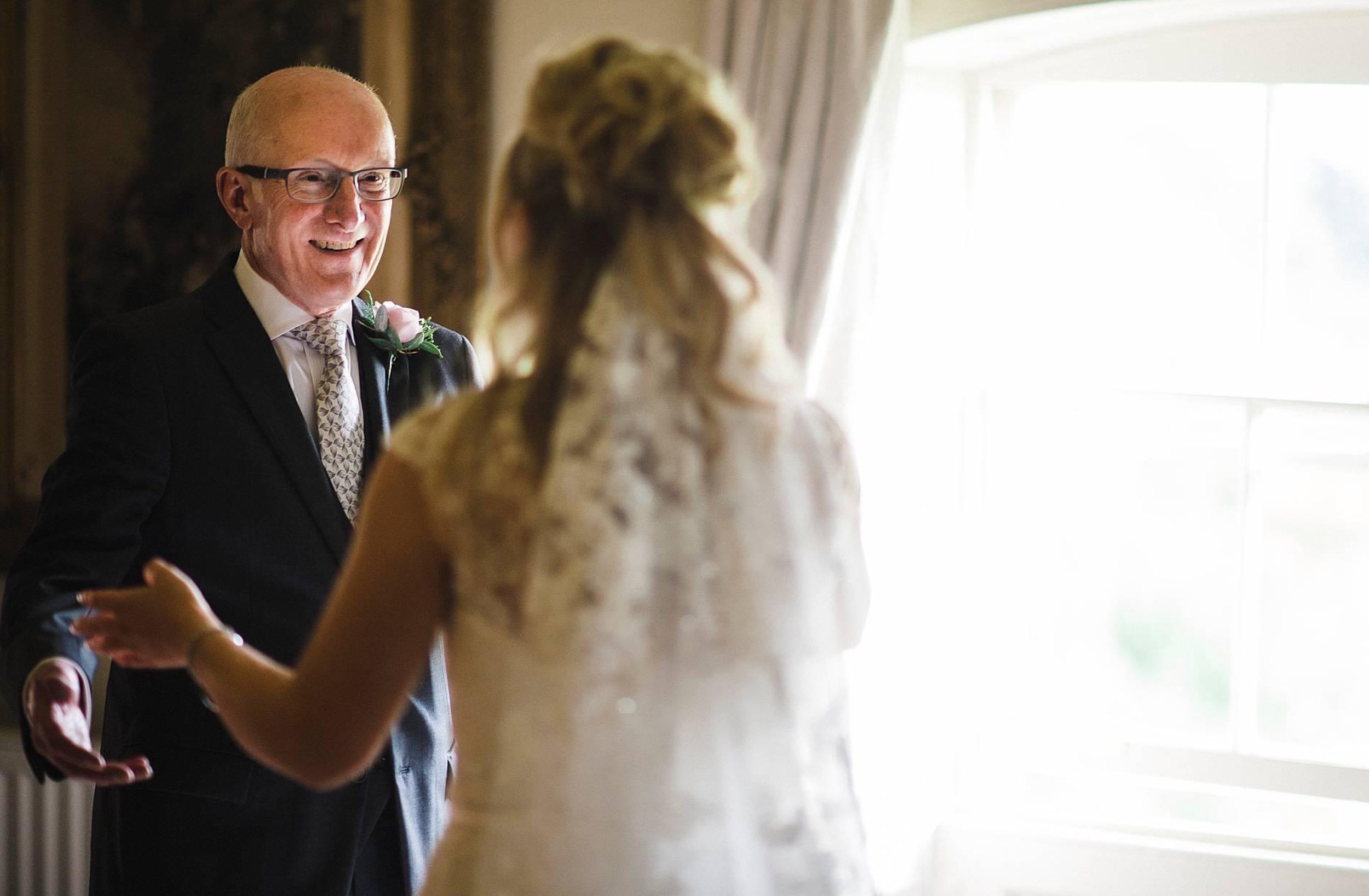 A man and woman share a moment indoors, with the man smiling and dressed in a suit, and the woman in a lace dress with her back to the camera, near a window with light coming through.