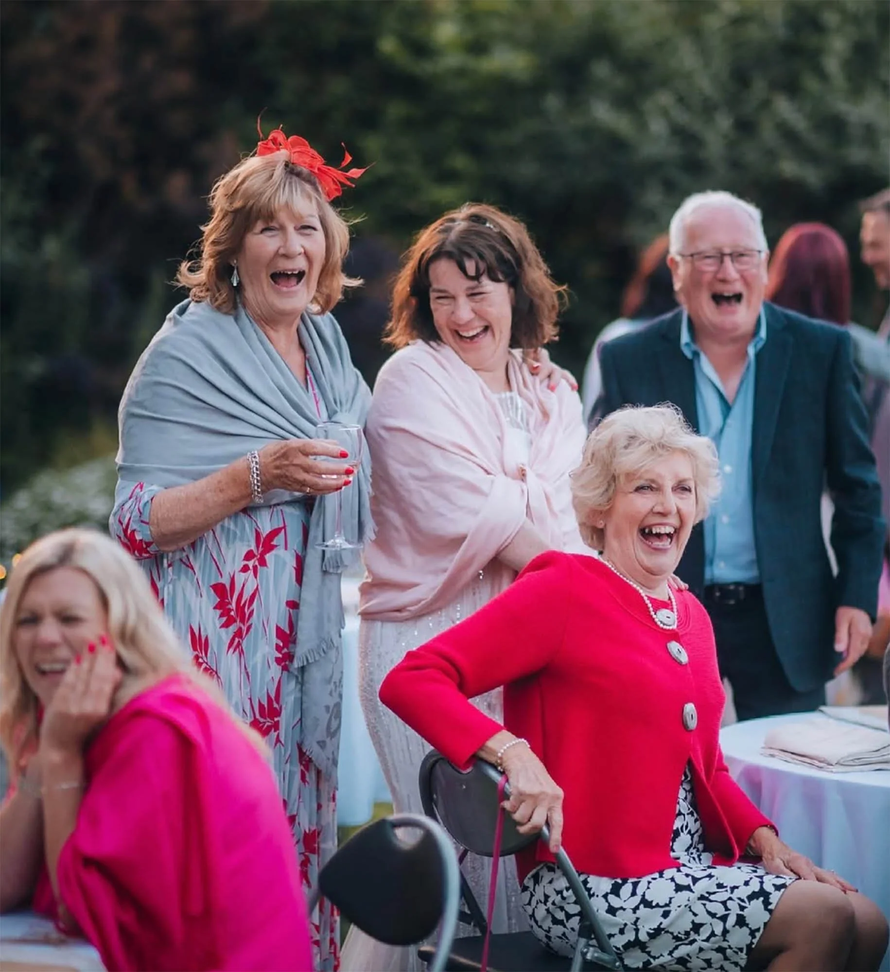 Group of laughing adults at an outdoor gathering, woman in red sitting on a chair, others standing behind and smiling.