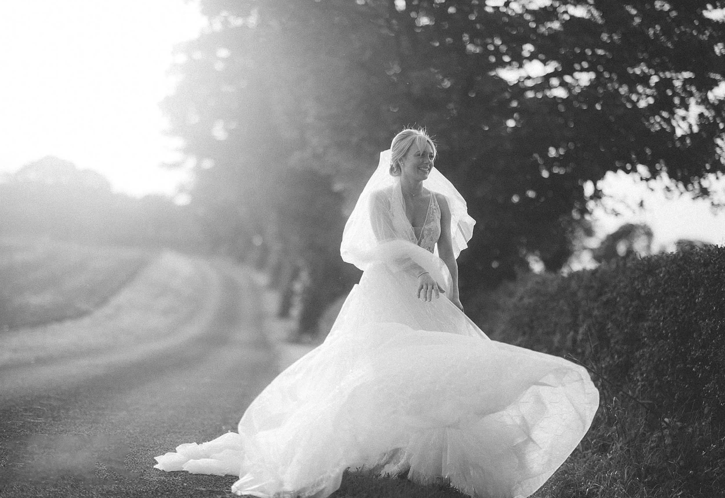 Black and white photo of a smiling woman in a wedding dress outdoors, standing on a dirt road near a hedge with trees in the background.