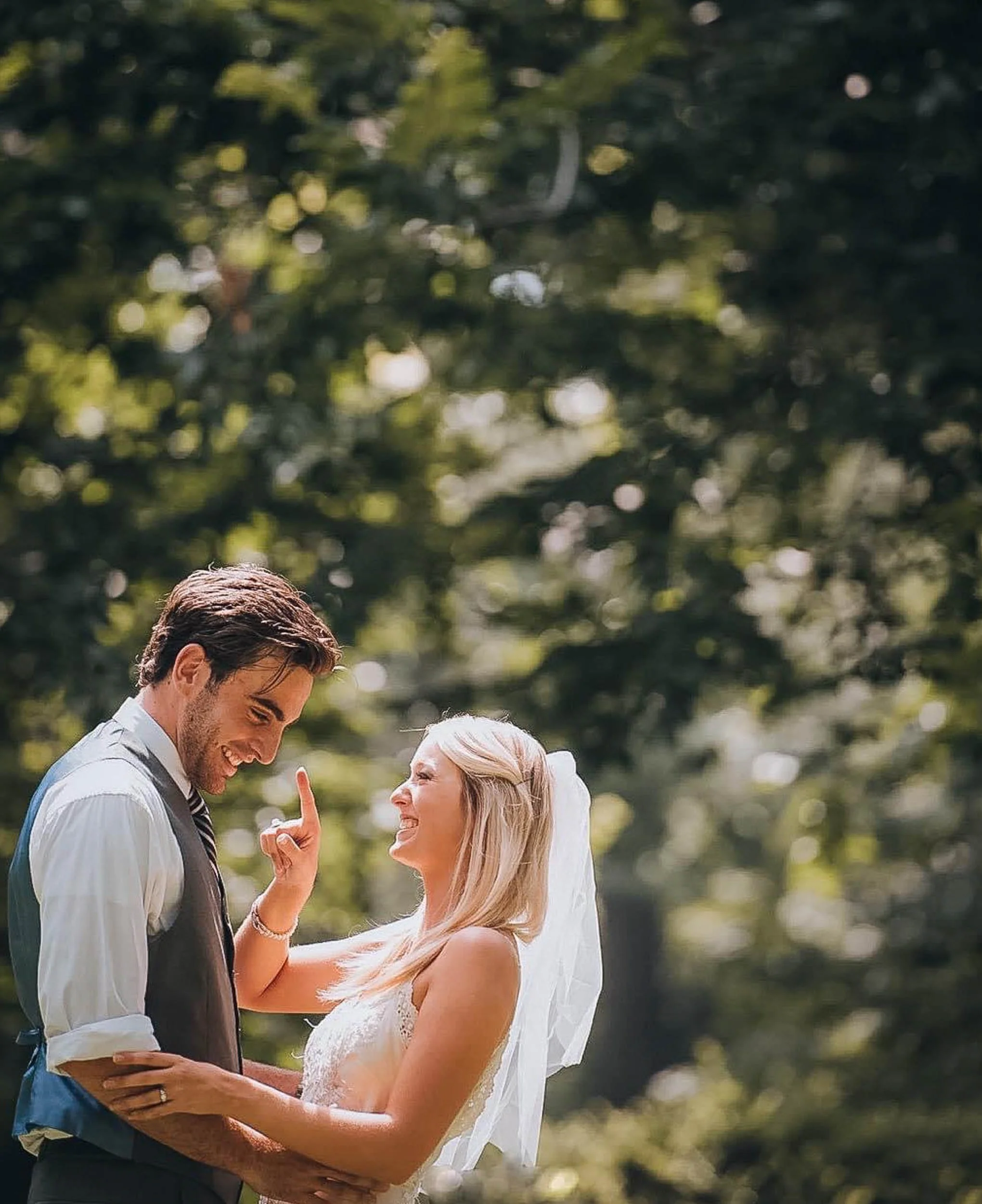 A happy bride and groom smiling and looking at each other outdoors during their wedding, surrounded by trees and greenery.