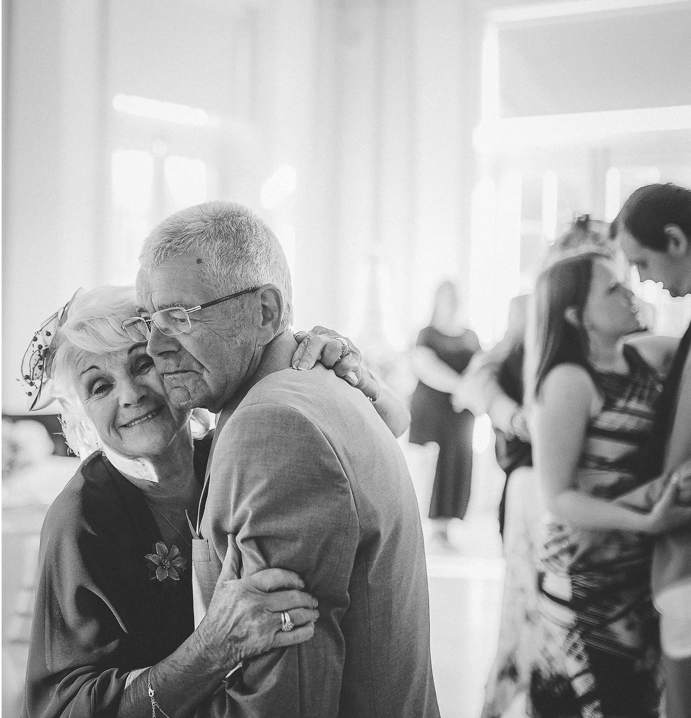 Black and white photo of an elderly woman and man dancing closely at a social gathering, smiling and enjoying each other's company while other couples dance in the background.