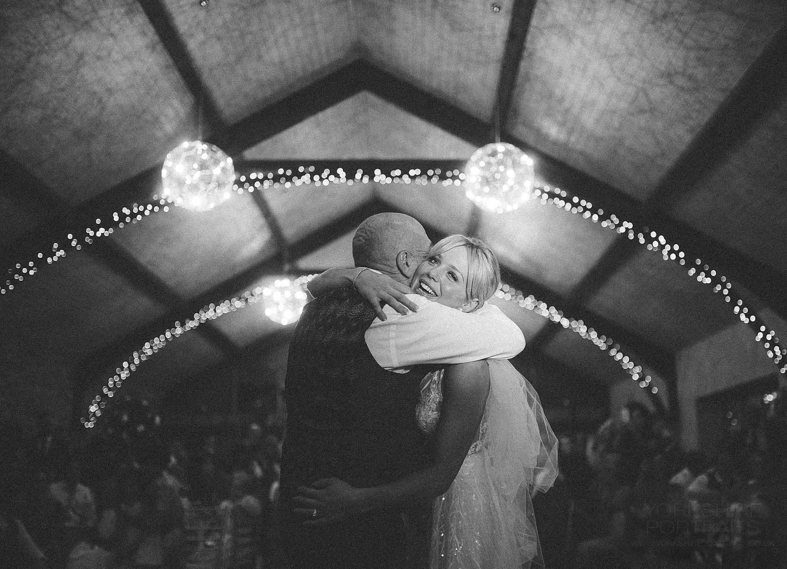 Black and white photo of a wedding dance, with a man and woman embracing and smiling. The venue is decorated with string lights and hanging spherical light fixtures, and there are blurred guests in the background.