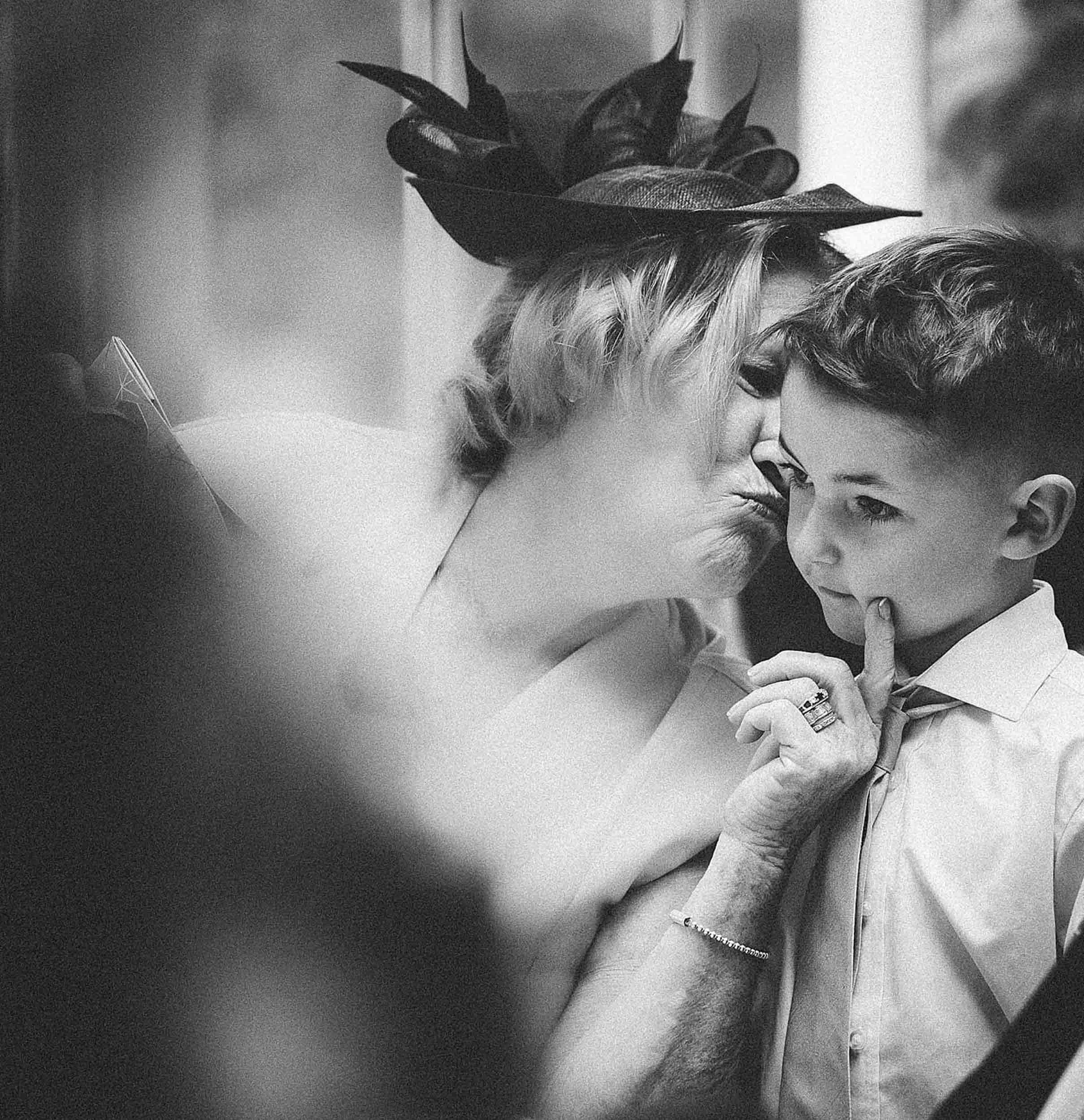 A woman with short wavy hair wearing a hat with leaves, touching forefinger to a young boy's chin, both with their noses close together in an intimate moment.