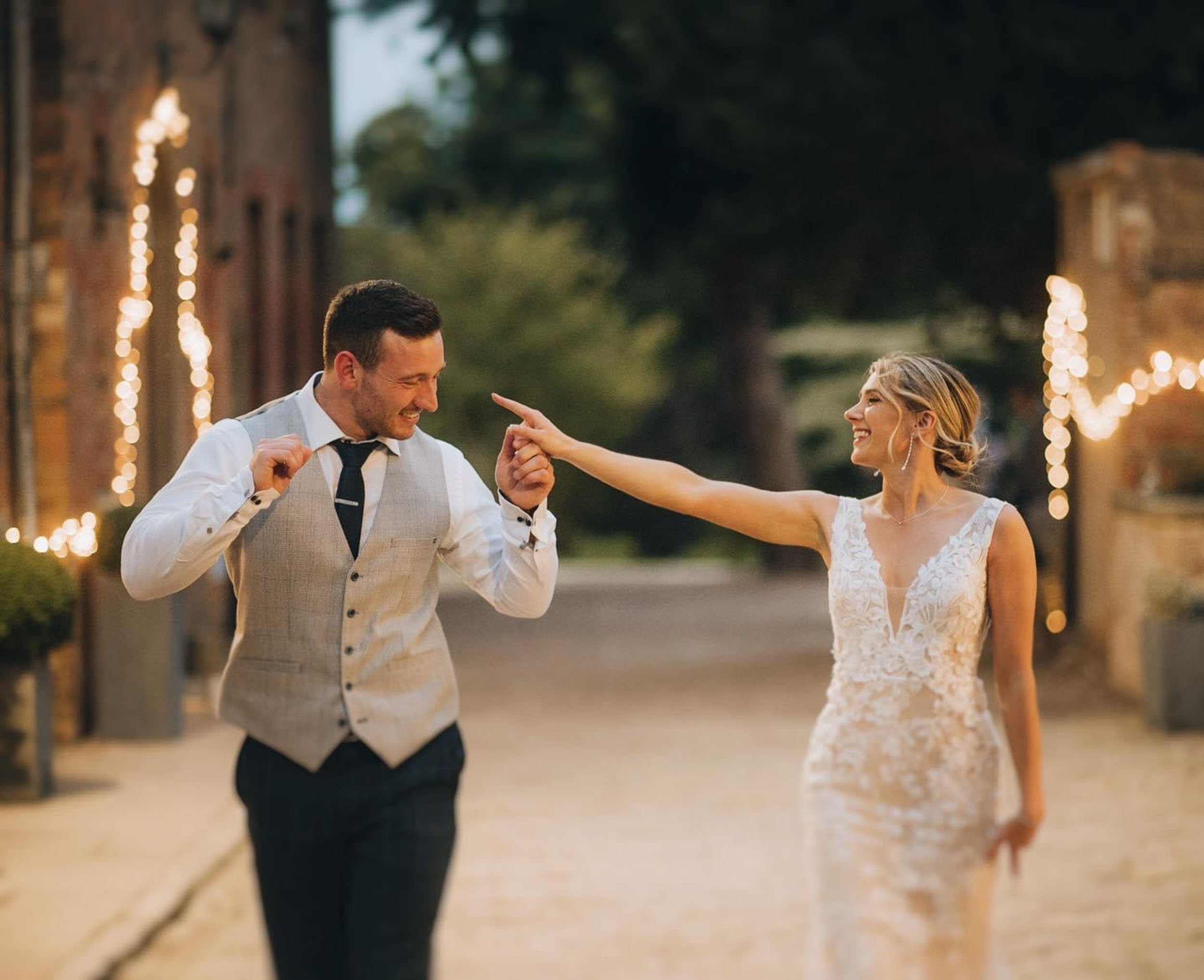 A bride and groom celebrate outdoors at sunset, with the bride playfully pointing at the groom. The groom is smiling, wearing a light gray vest and dark pants. The bride is smiling and in a white lace wedding dress. Decorative string lights are visib