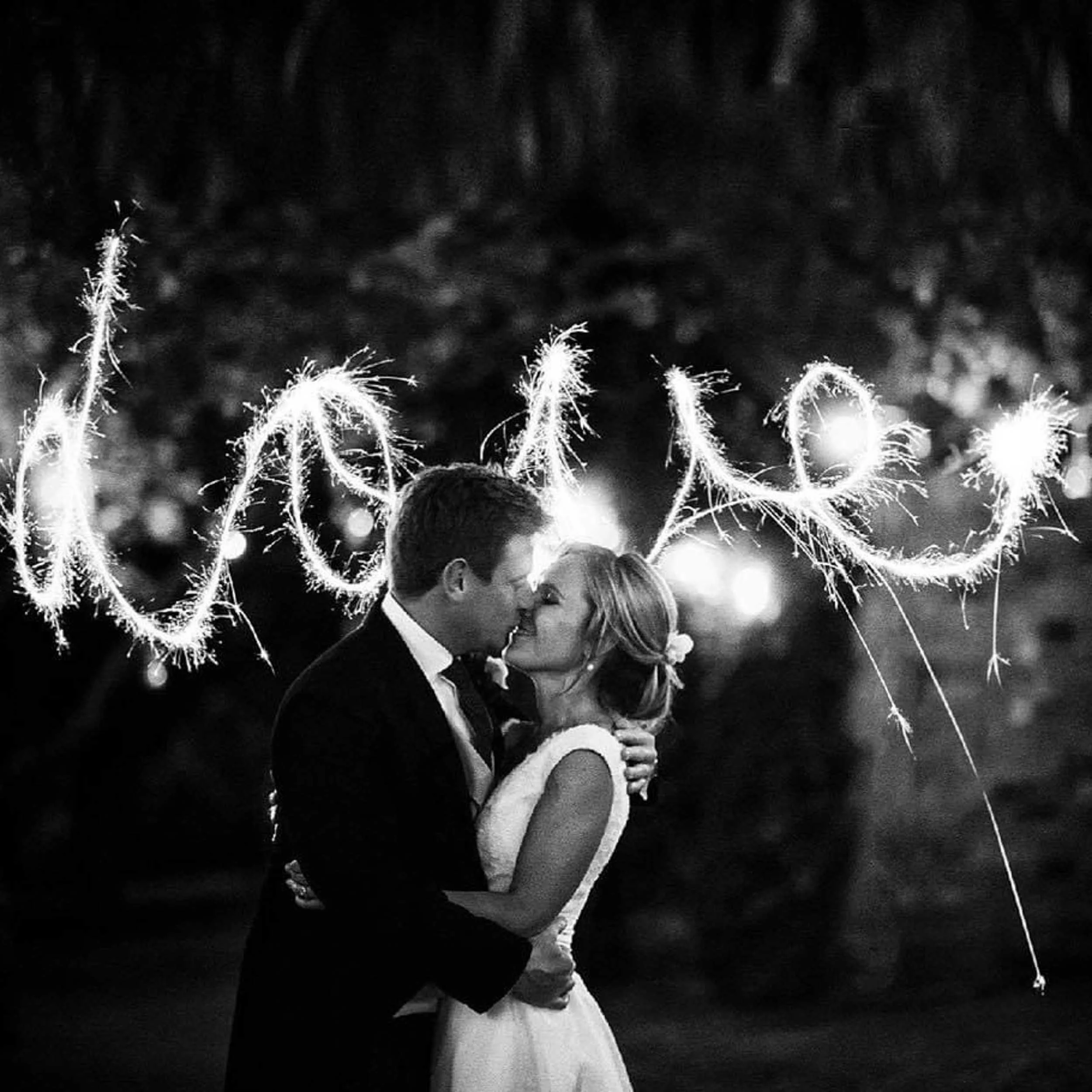 A black and white photo of a couple in wedding attire sharing a kiss at night, with sparklers forming the word 'love' in the background.