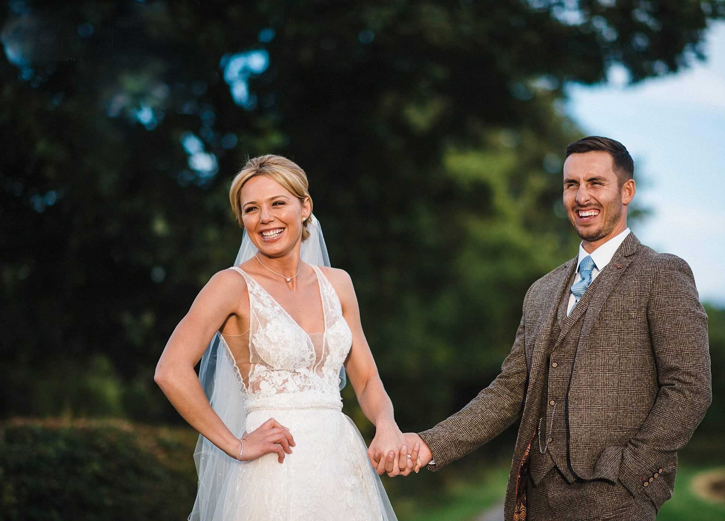 A bride and groom hold hands outdoors, smiling, with trees in the background.