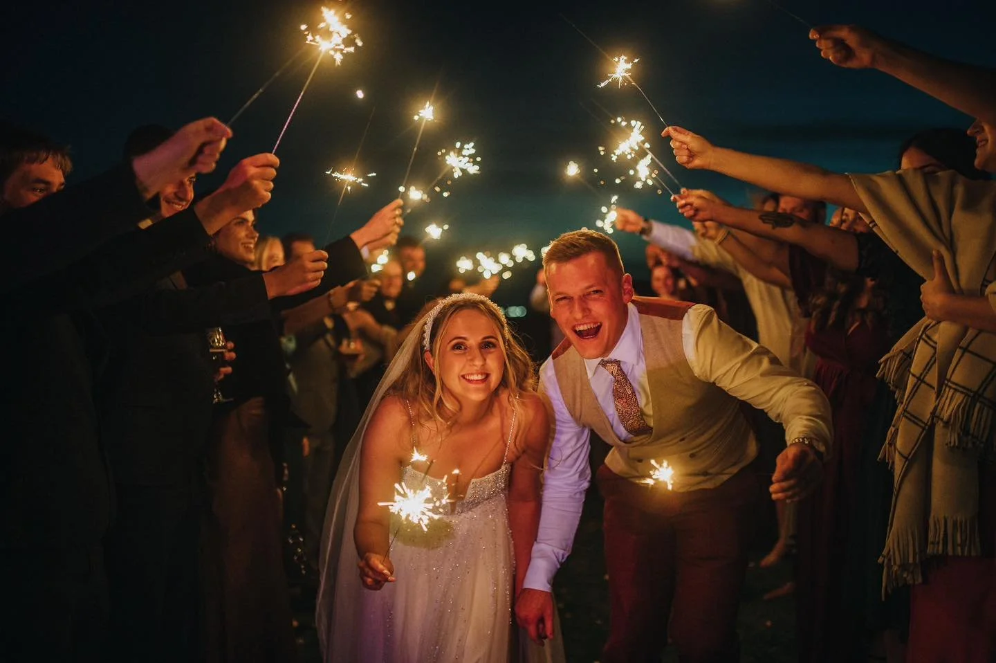 A group of people holding sparklers at night, celebrating a wedding with the bride and groom smiling in the center.