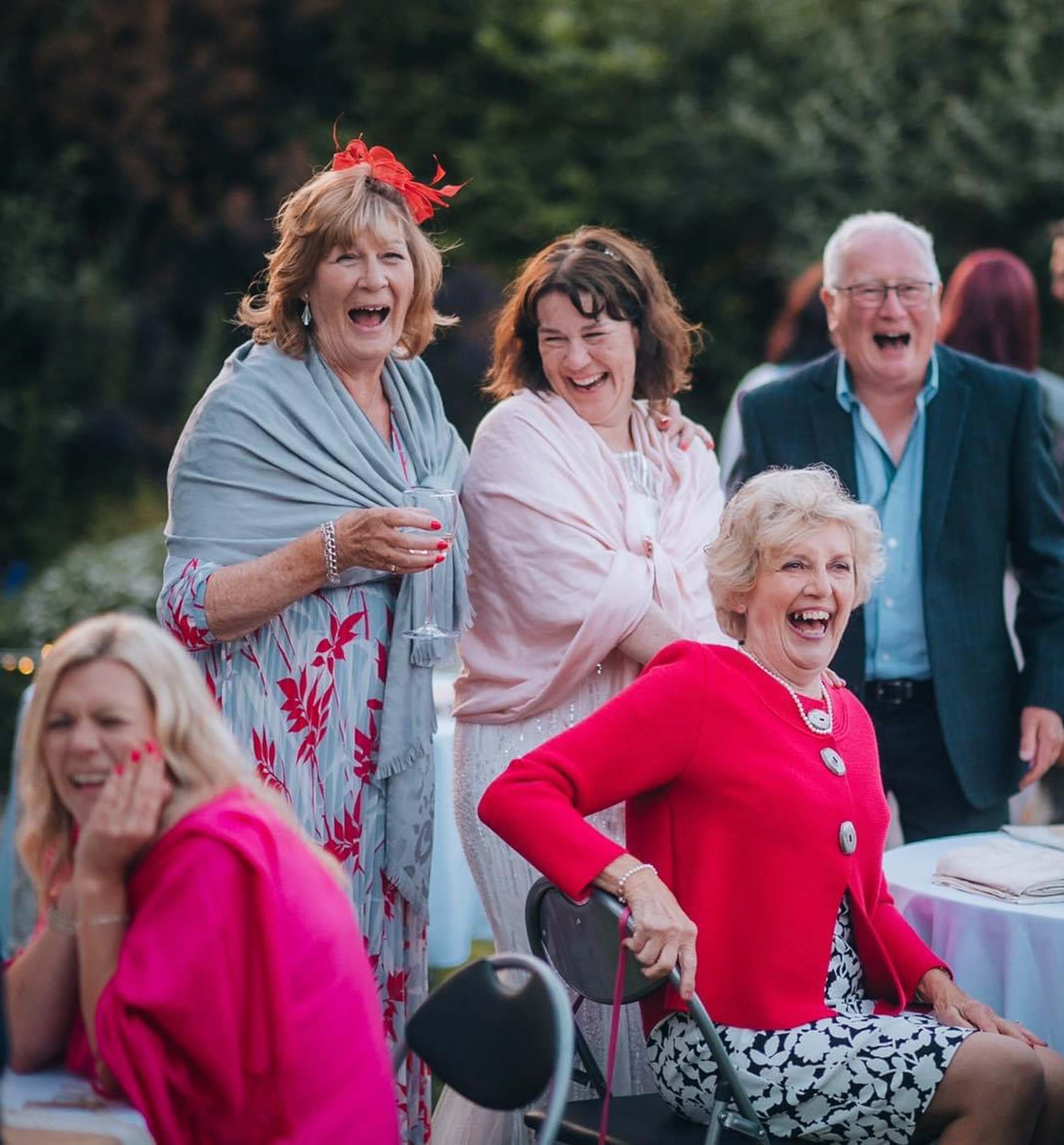 Group of elderly women and a man at a celebration outdoors, laughing and smiling, dressed in colorful clothing, with tables and greenery in the background.