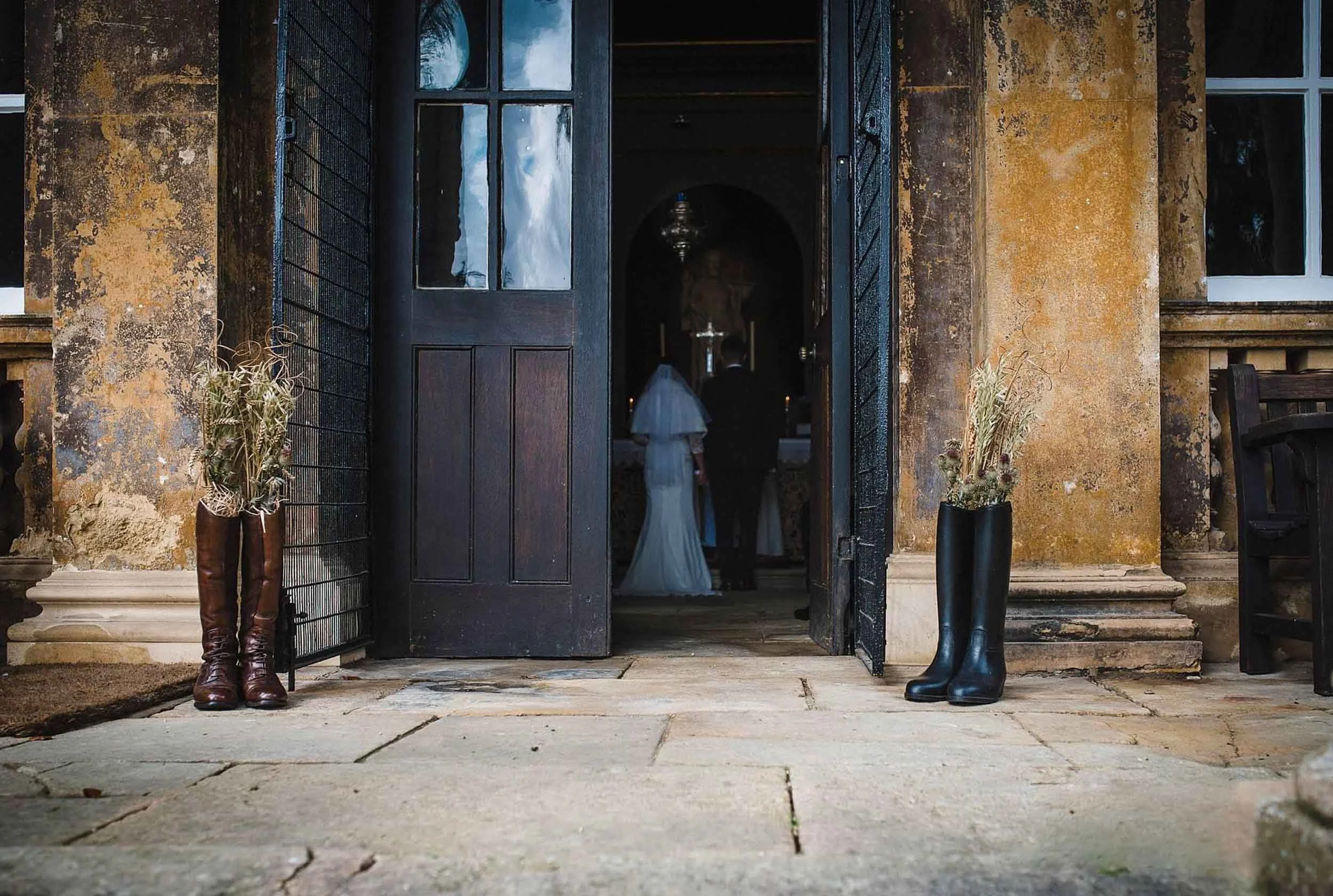 Open doorway of an old stone building with two pairs of tall boots filled with dried flowers on either side. Inside, a bride in a white wedding dress and veil stands with a groom in a black suit near an altar.