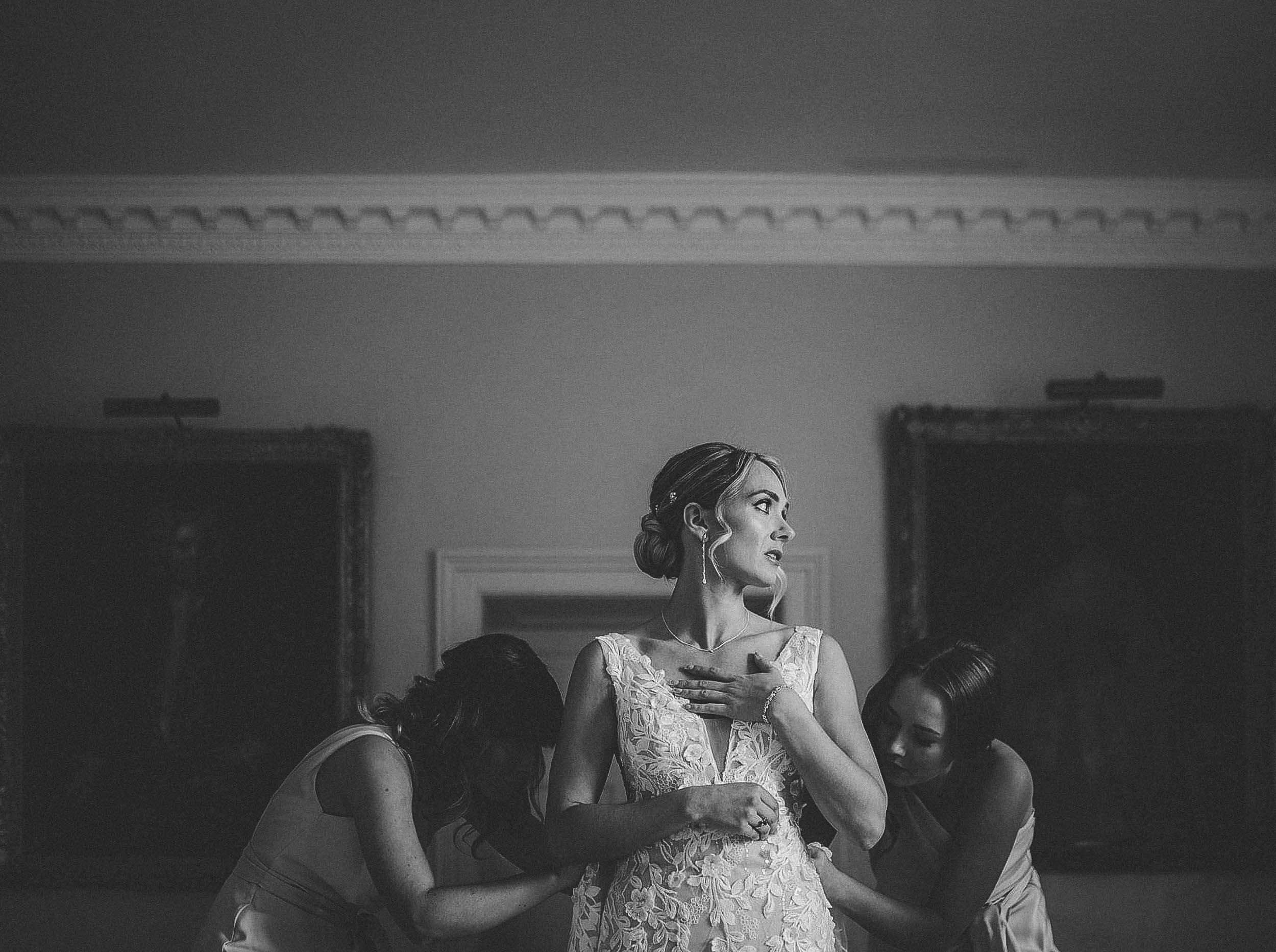Black and white photo of a bride in a lace wedding gown being assisted by two women, with the bride placing her hand on her chest and looking to the side.