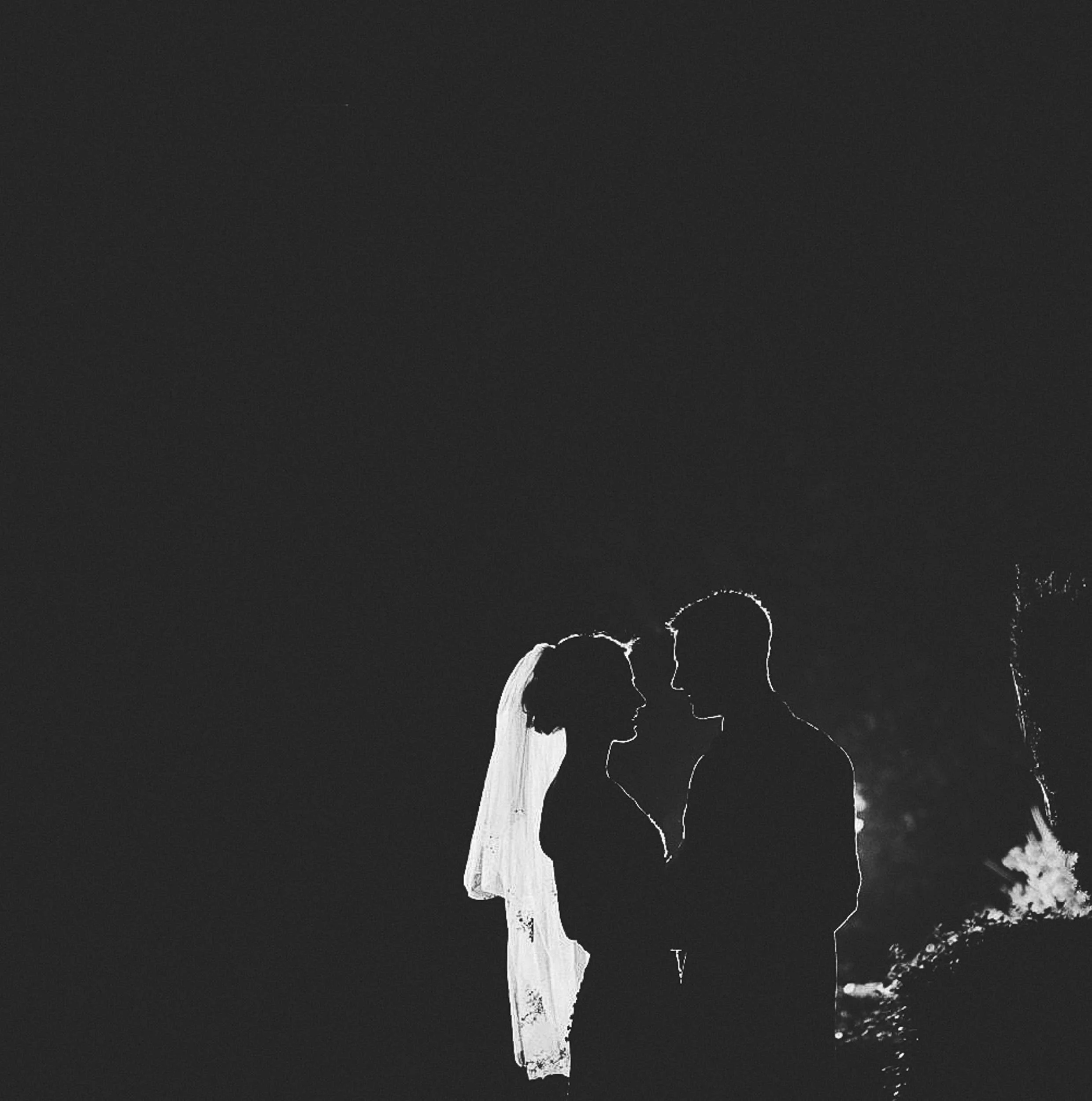 Silhouette of a bride and groom facing each other at night, with a fireworks display in the background.