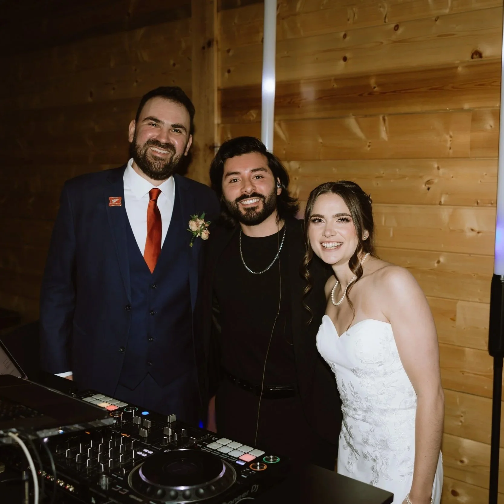Three people smiling at a wedding reception, standing behind DJ equipment with a wooden wall in the background. The man on the left is wearing a dark suit with a white shirt, red tie, and boutonniere. The man in the middle has dark hair, a beard, and is wearing a black jacket with a chain necklace. The woman on the right is wearing a strapless white dress with pearl jewelry.