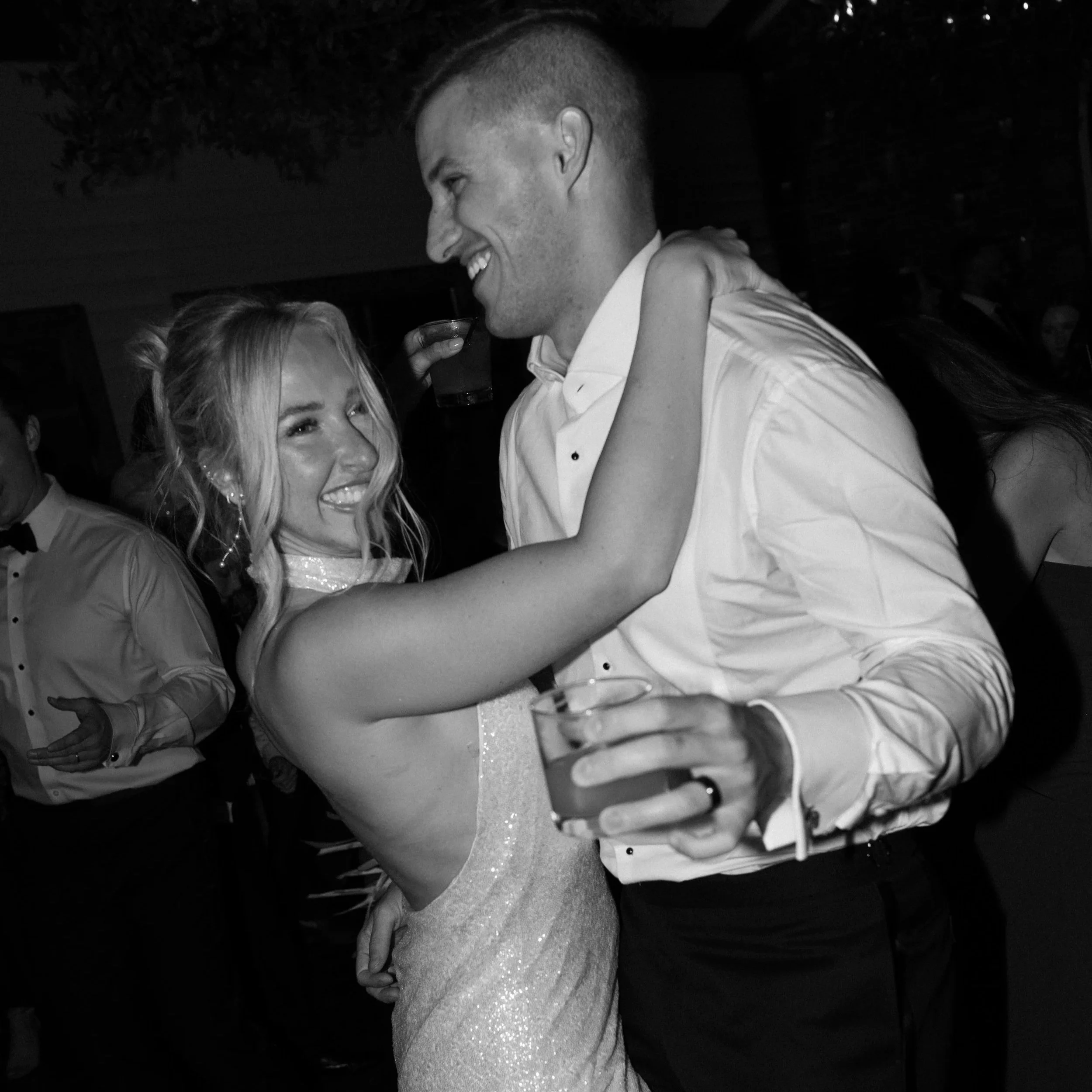 A couple is dancing closely at a formal event, smiling and holding drinks, with other people in the background.