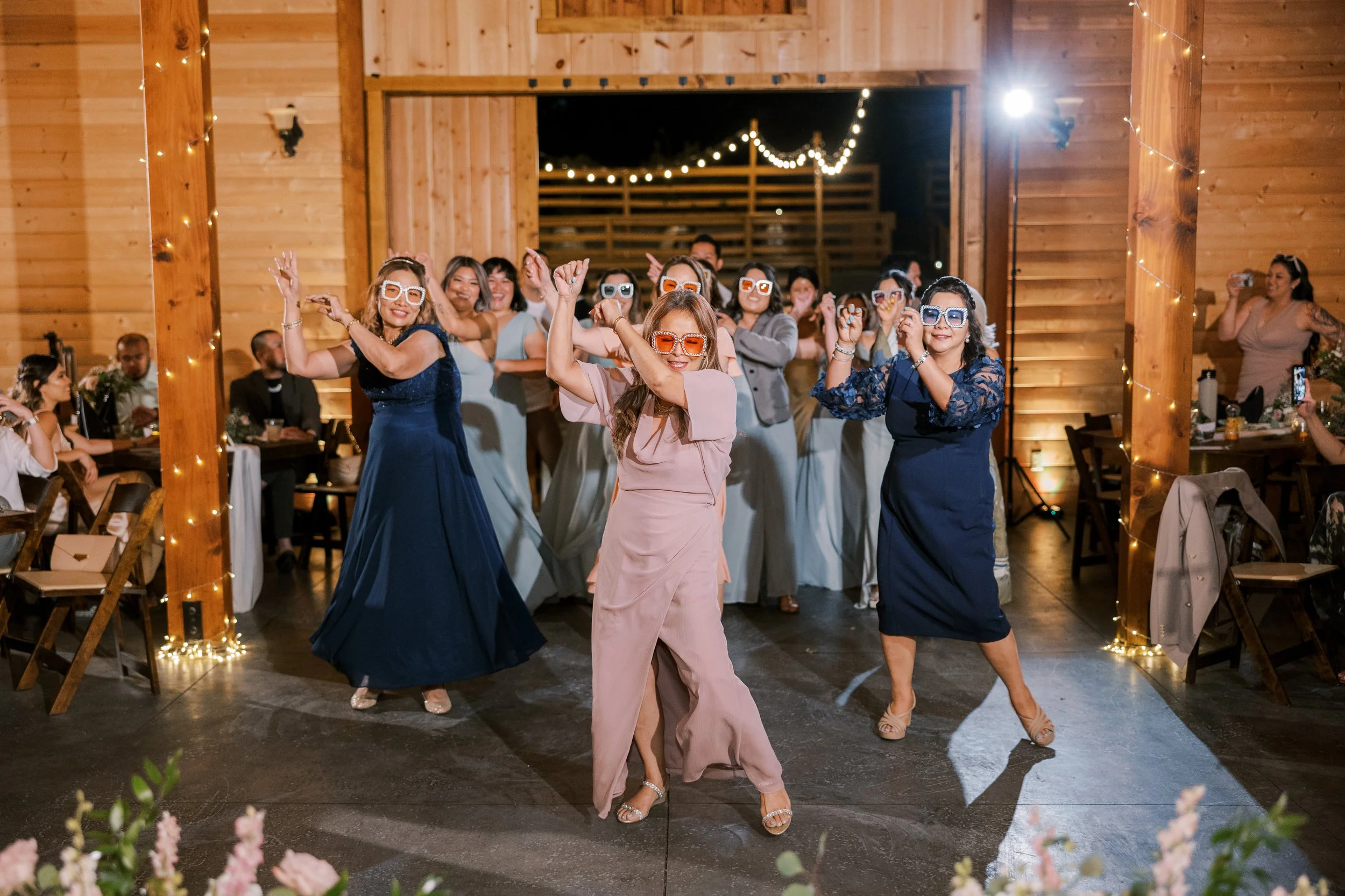 A group of women dancing and enjoying themselves at a celebration in a wooden venue, with string lights overhead and guests seated at tables.