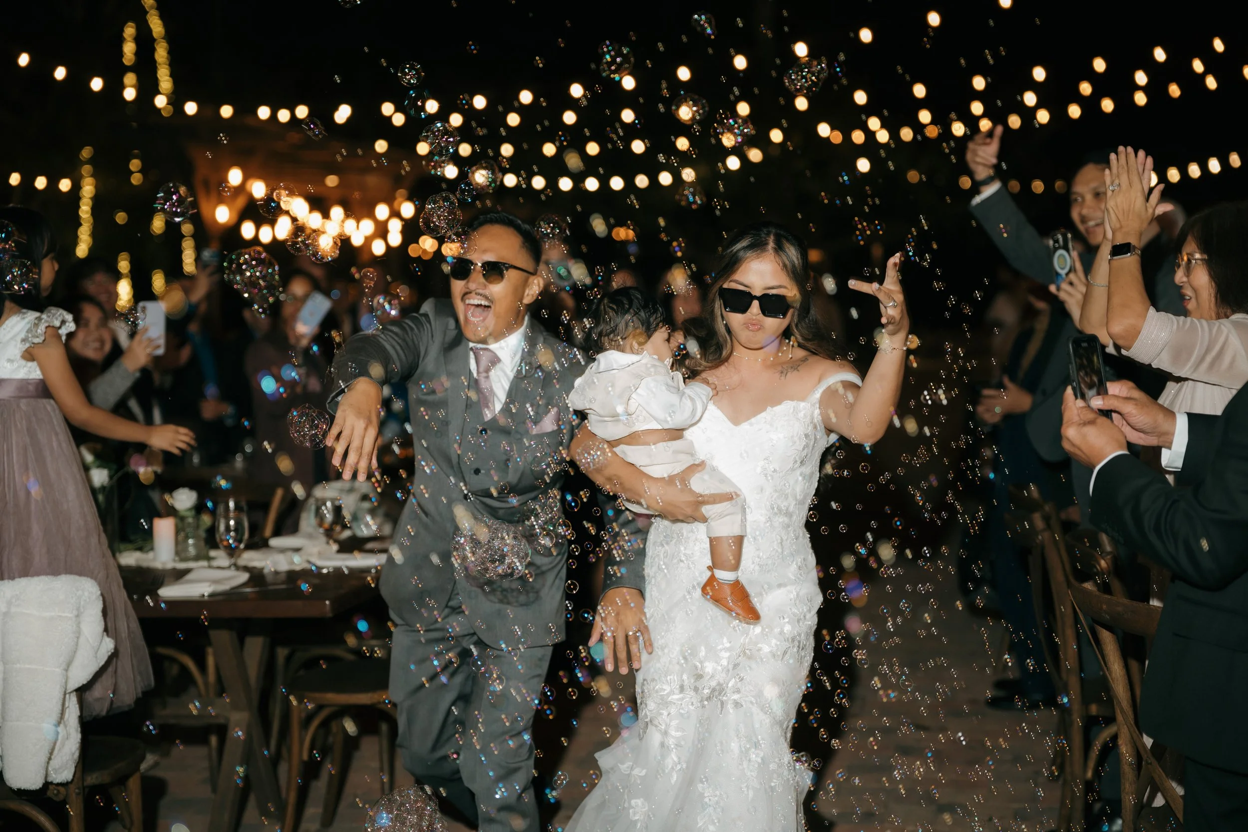 A bride and groom wearing sunglasses celebrating at their wedding reception, surrounded by guests taking photos, with bubbles and string lights overhead.