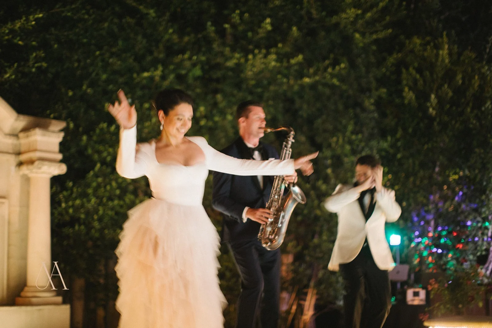 A woman in a white dress dancing with a man playing a saxophone at an outdoor event at night, with another man standing in the background near colorful lights.
