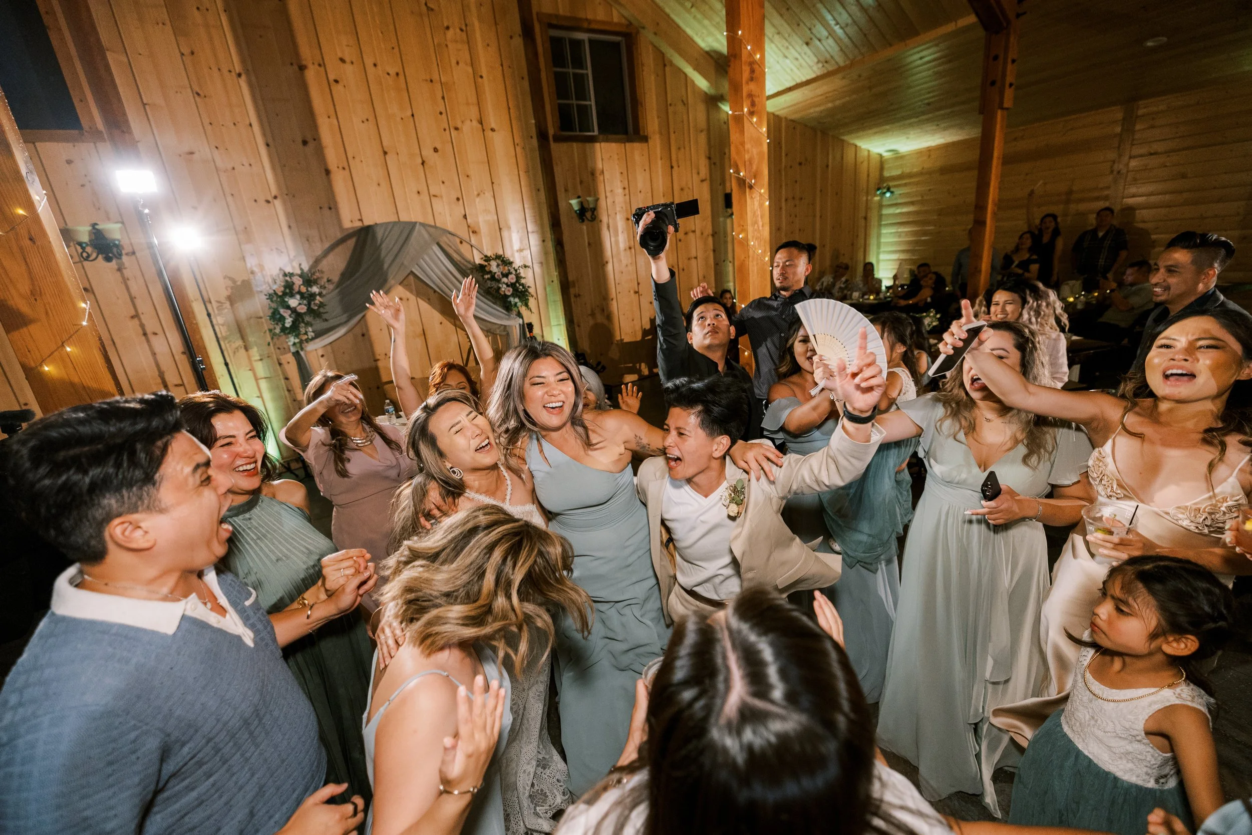 Group of people dancing and celebrating at a wedding reception inside a wooden hall with decorations and string lights.