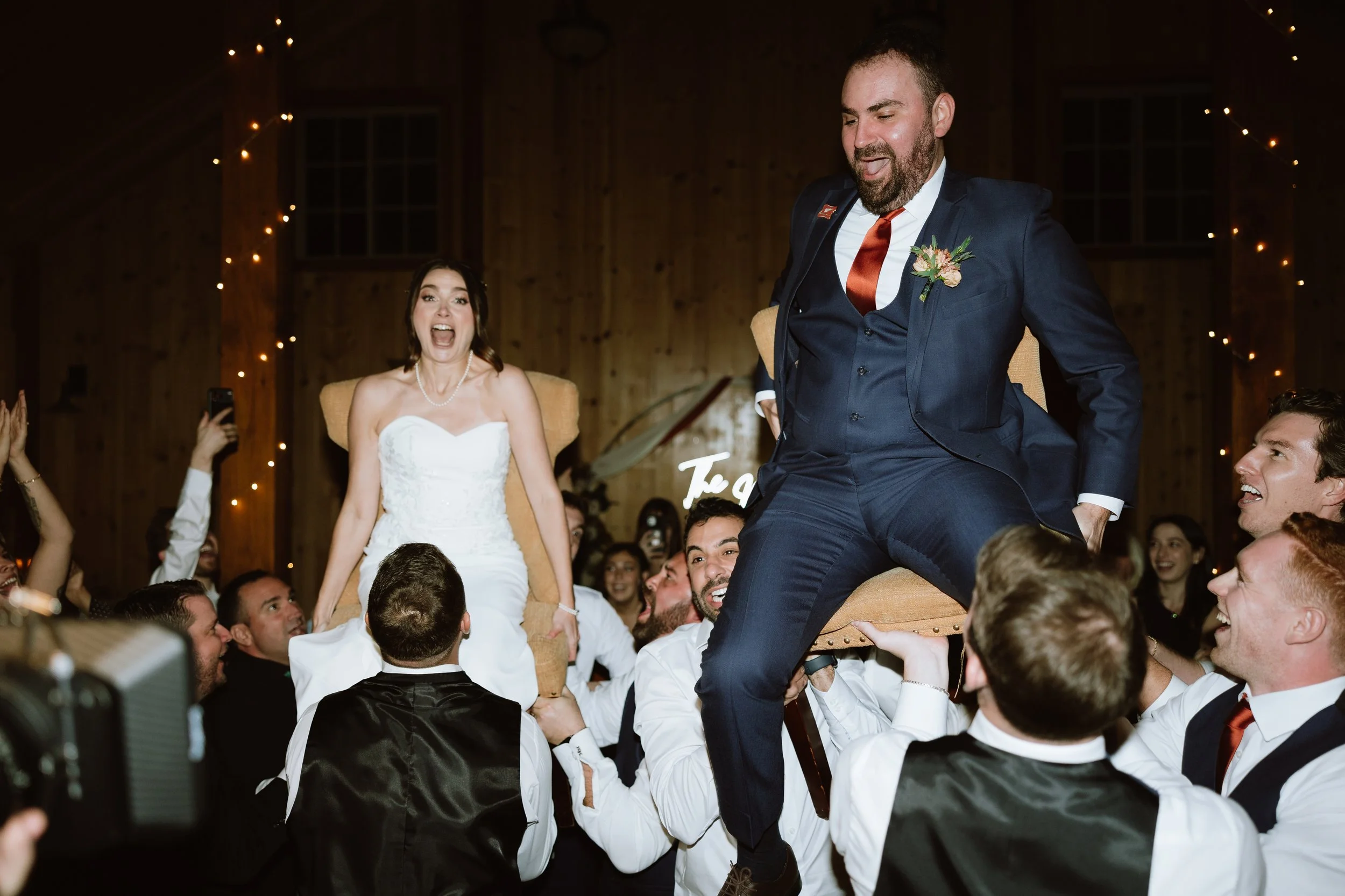 A wedding reception scene with a bride in a white wedding dress sitting on a chair held by friends, and a groom in a navy suit sitting on the chair with his legs crossed, smiling. The background features wood-paneled walls with string lights, and gue