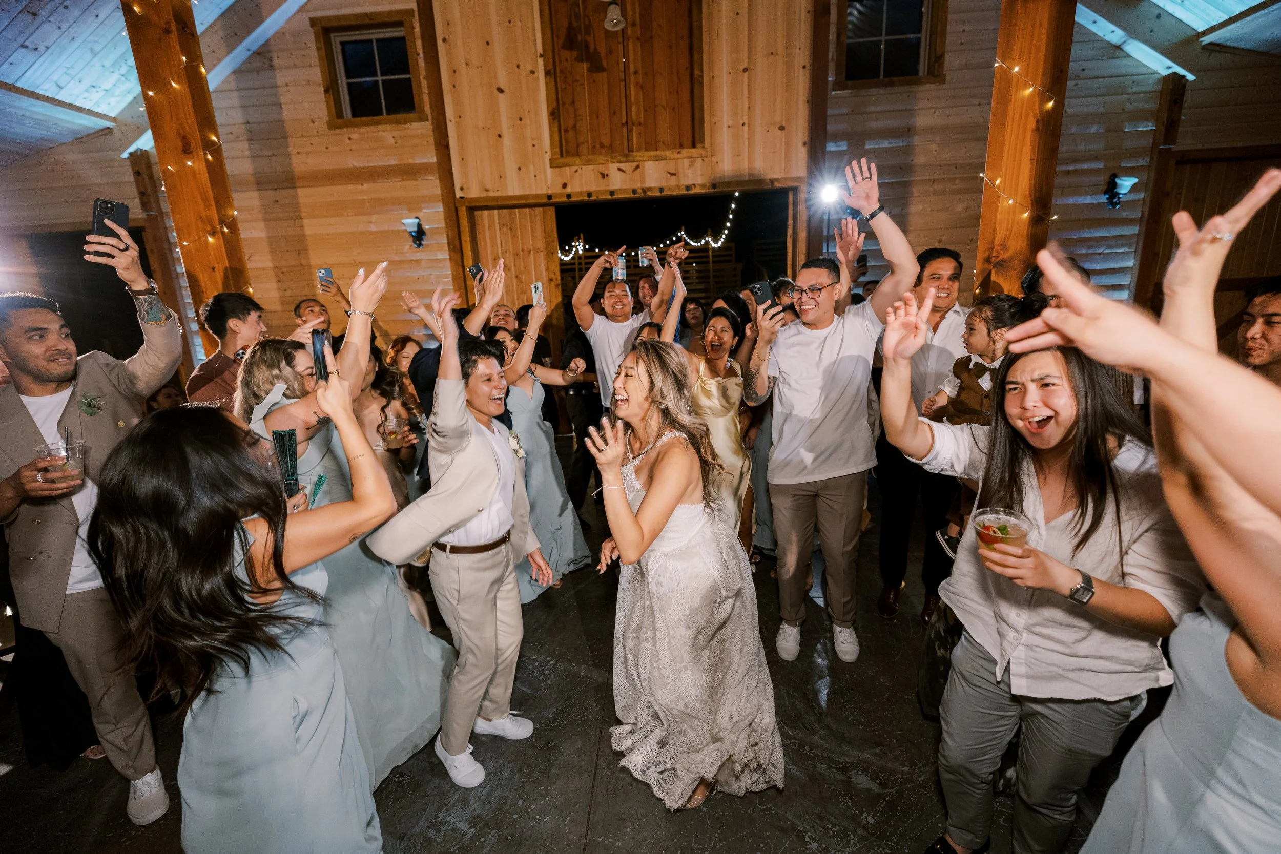 People dancing and celebrating at a wedding reception in a wooden venue, with some holding drinks and taking photos, the bride in a white dress smiling in the center.