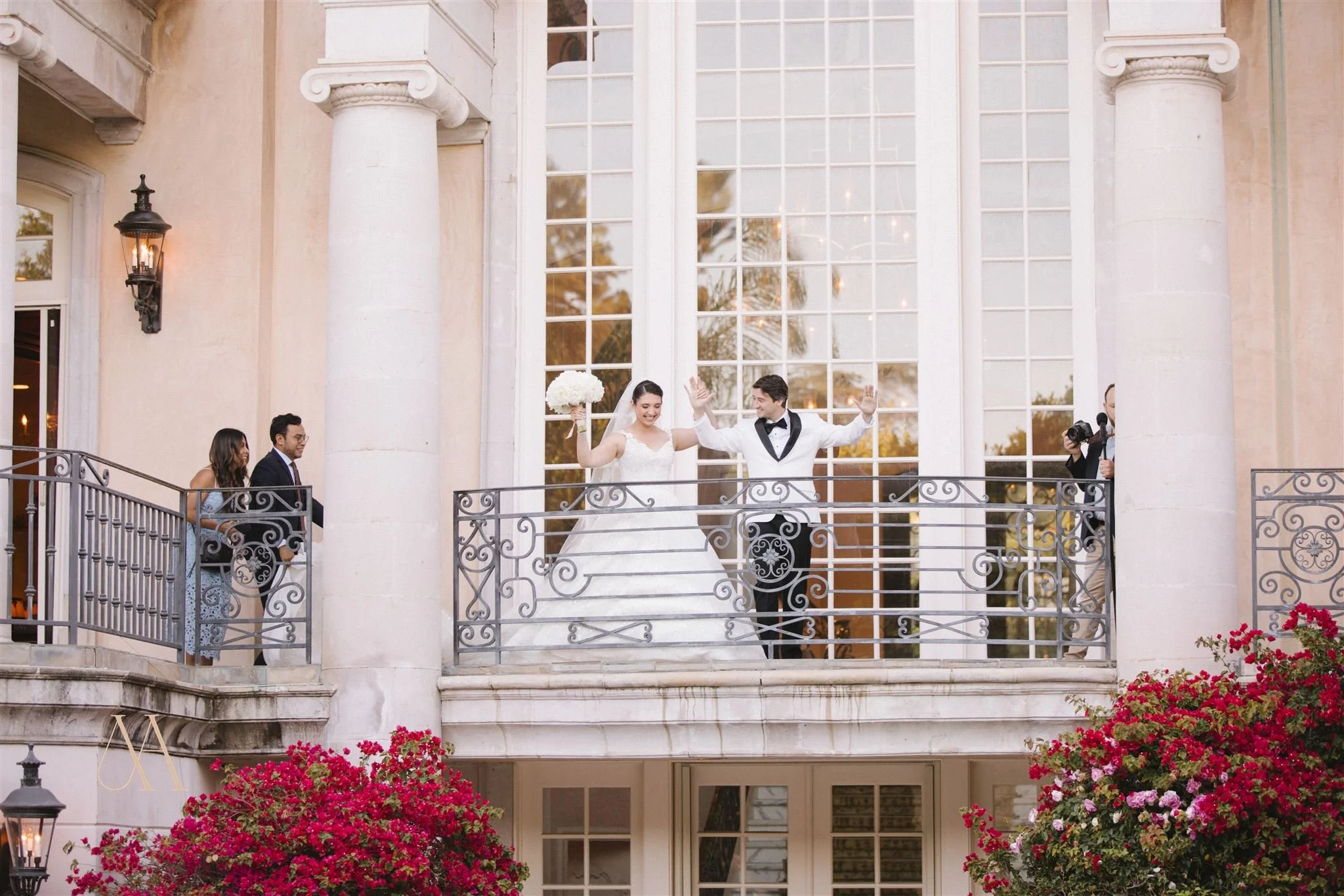 Bride and groom celebrating on a balcony during their wedding, with guests and a photographer nearby, pink flowers in foreground, large window and columns in background.