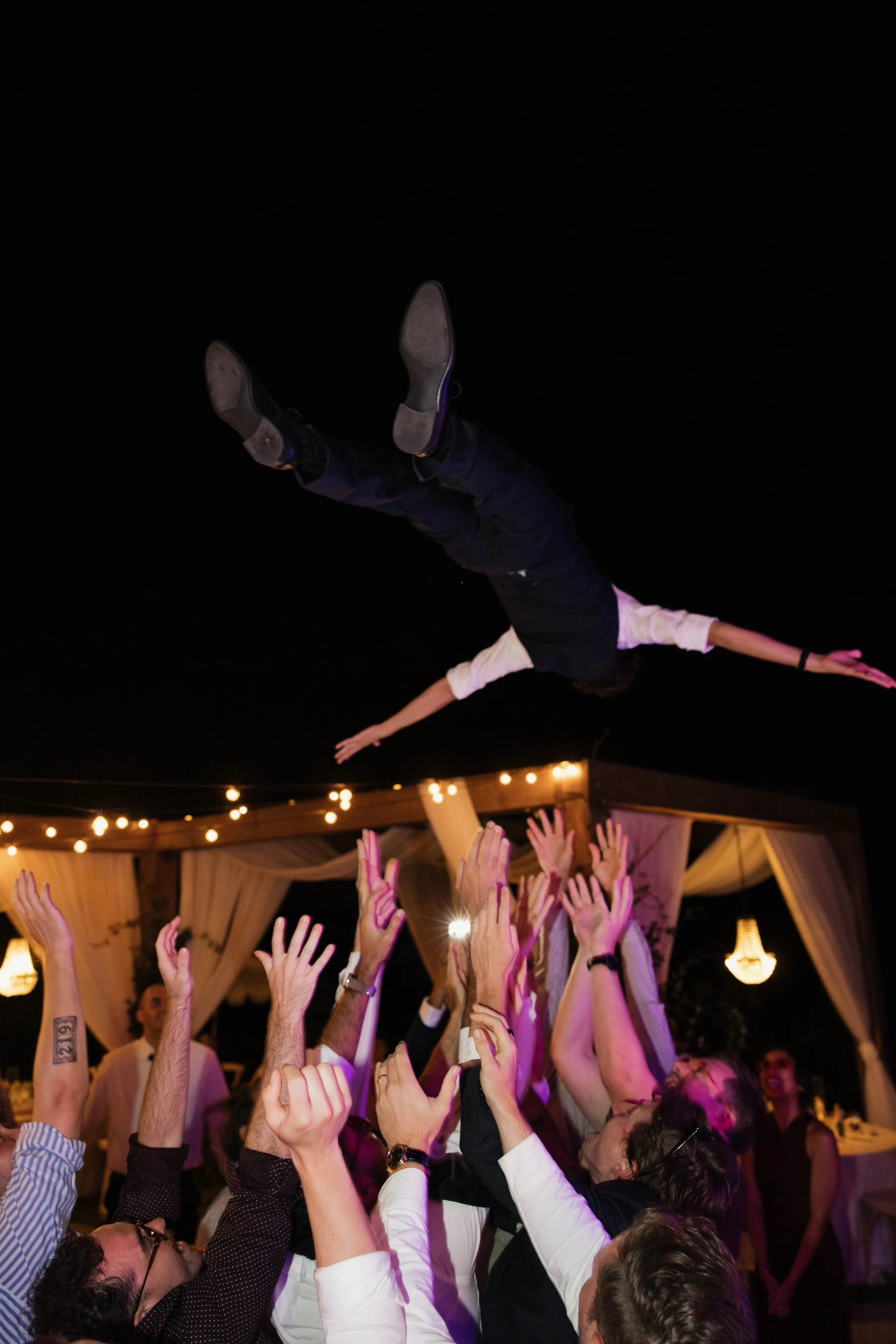 Person being tossed in the air at a celebration or party with string lights and a decorated tent in the background at night.