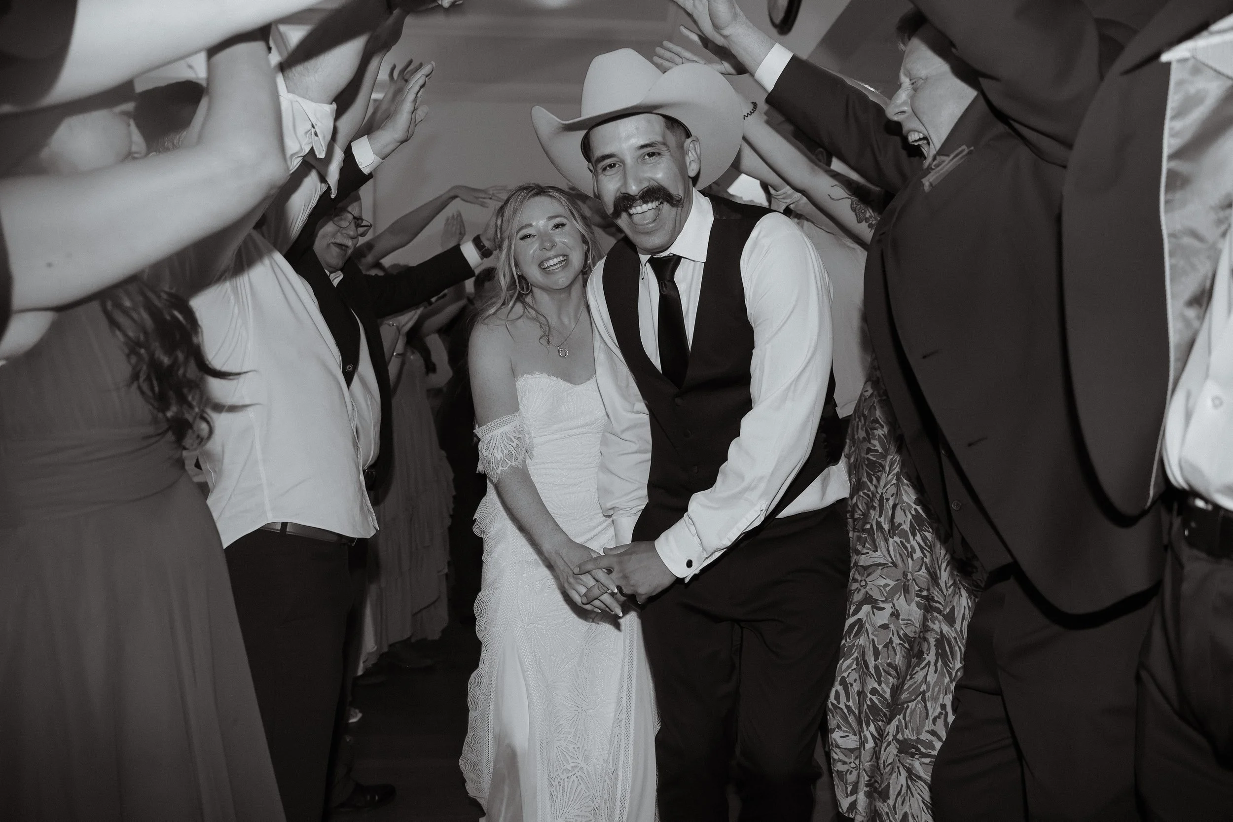 A couple holding hands and smiling, walking through a tunnel of people raising their arms, at a wedding reception, in black and white.